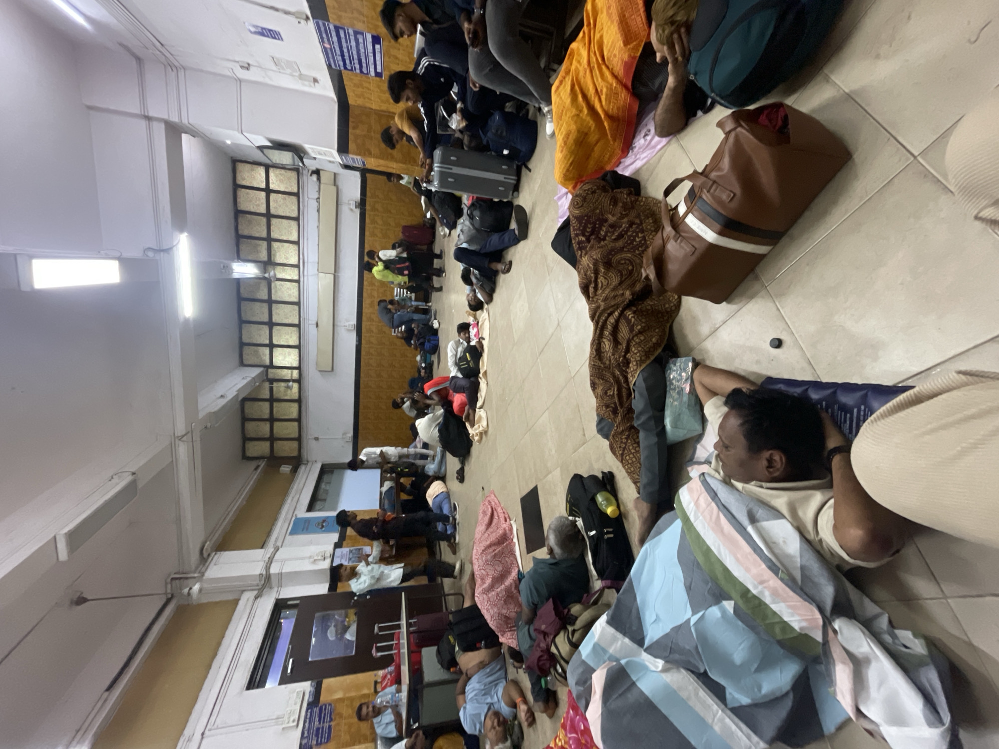 In a crowded waiting room, likely at a train station in Mumbai, India, a multitude of people are waiting. The scene is dominated by individuals resting on the floor, some using blankets, bags, or luggage as makeshift pillows or mattresses. Several individuals are seated on benches along the walls. Various pieces of luggage, including suitcases and bags, are scattered throughout the room. The waiting area is equipped with windows, doors, and overhead lighting. Signage can be seen on the walls. The tiled floor shows signs of wear and tear.