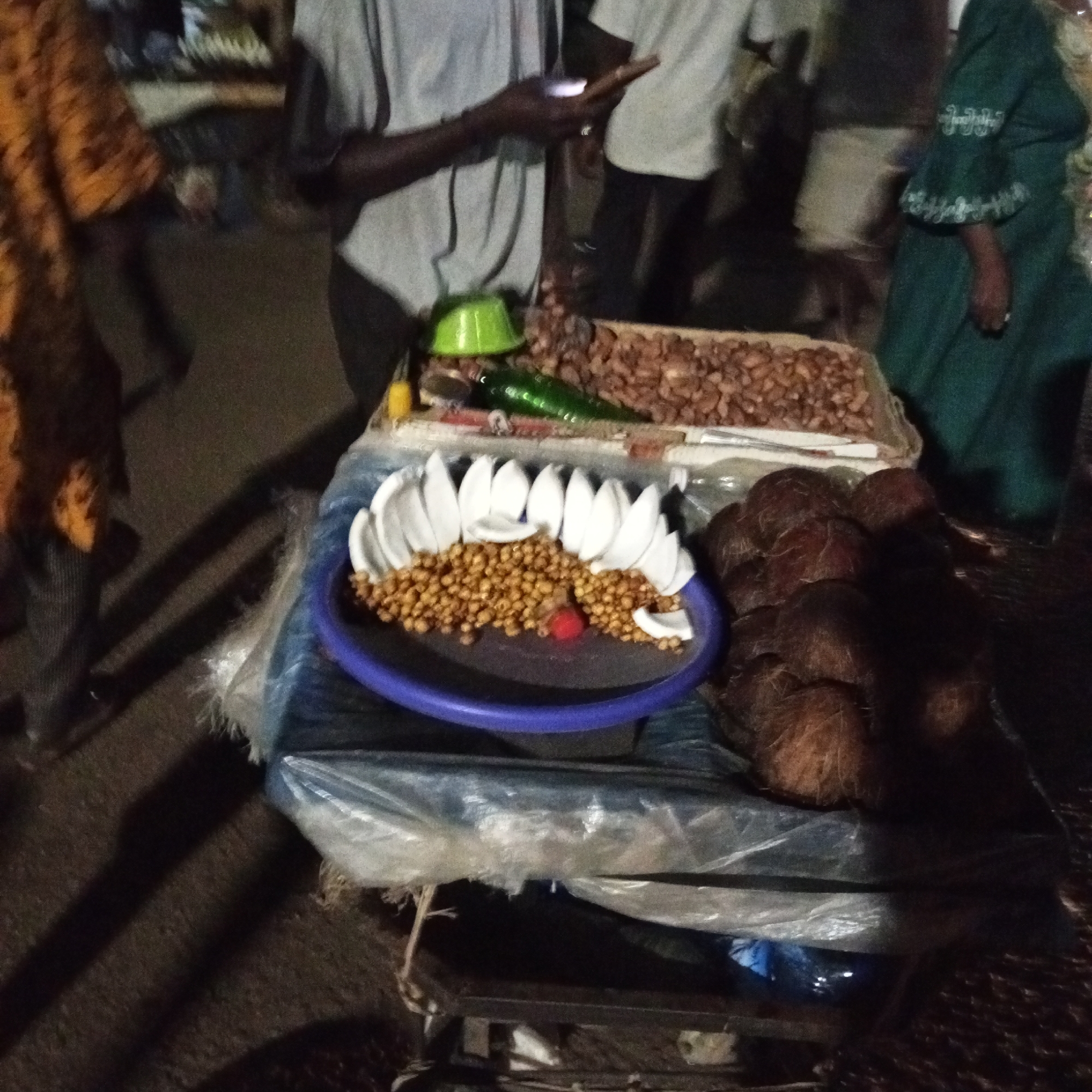 The image shows a street vendor's cart in an unidentified location in Unknown, Unknown. The cart, draped in a clear plastic sheet, displays various items for sale. In the foreground, a blue plastic plate holds a ring of white coconut pieces and what appears to be roasted chickpeas. Several whole coconuts are stacked next to the plate. A tray behind the plate contains more goods, including roasted nuts, green peppers, and a small green cup. Several individuals are visible around the cart. One person is standing behind the cart using a mobile phone. Another person wearing a green outfit is partially visible on the right side of the image. Other people are walking in the background. The environment appears to be dark, suggesting the scene is set at night.
