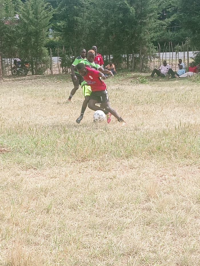 The image depicts an outdoor football match in Lwandeti ward, Kenya. In the foreground, two male individuals are actively engaged with a white football on a dry, sparsely green grass field. One player, wearing a red short-sleeved jersey with black trim, black shorts, and black athletic shoes, is in the process of dribbling the ball, leaning forward with his left foot on the ball and his right leg extended. The second player, wearing a lime green short-sleeved jersey, lime green shorts, and black athletic shoes, is positioned closely behind the first player, appearing to challenge for the ball. His hair is dyed a light blonde or yellow color.
In the mid-ground, other individuals are visible. Two men stand behind the active players; one in a green shirt and another in a red shirt. Further back, along the edge of the field, approximately six to eight individuals are seated or lounging on the ground, observing the match. They are dressed in casual attire, including shirts and trousers of various colors.
The background features a line of tall, leafy green trees, suggesting a forested or treed area. A wooden post fence runs along the perimeter of the field, visible between the spectators and the trees. To the far left, a motorcycle with a dark seat and light-colored frame is parked near the fence. The sky is bright, indicating daylight conditions.