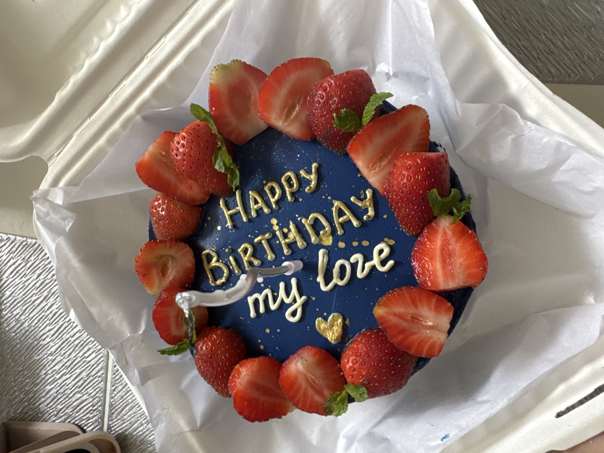 The image shows a round birthday cake in a white take-out container. The cake has a blue frosting decorated with gold lettering that says "Happy Birthday my love," along with white cursive writing. The cake is also decorated with sliced strawberries placed around the edge and sprigs of mint. The cake rests on white tissue paper within the container. The location in the city of Unknown, Unknown is unknown.