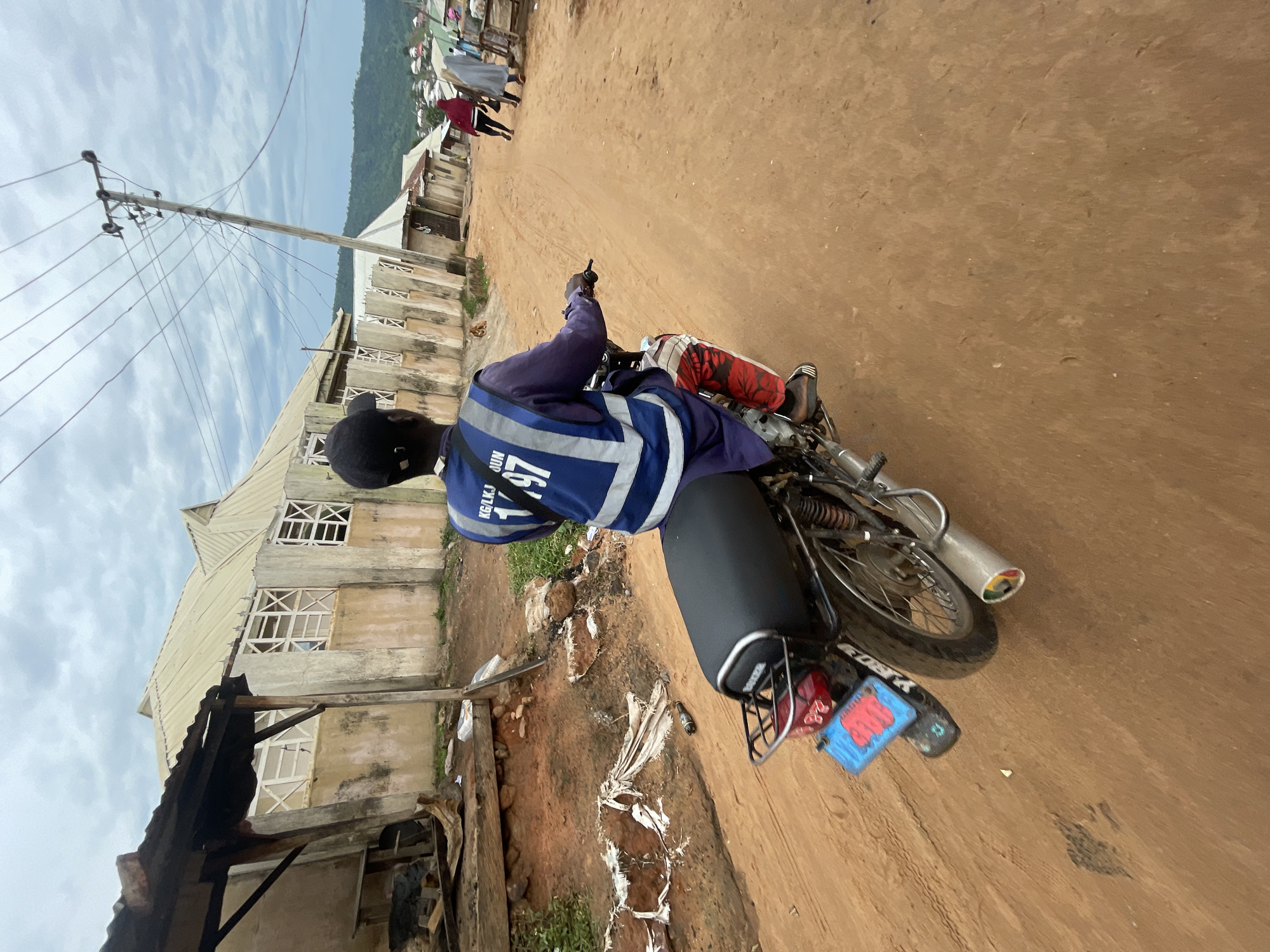 A man wearing a blue vest with reflective stripes and "KG LK/KJ JUN 1/97" printed on it rides a motorcycle on a dirt road. He has a black cap on and is wearing red and white patterned pants. The motorcycle has a blue license plate with red lettering. In the background, there are buildings with light-colored walls and light-colored roofs. There is a utility pole with multiple wires running across the scene. Two people, one in a red garment and another in a light-colored garment, are walking away from the viewer in the distance. The ground is sandy and appears dry. The sky is cloudy. The scene appears to be in the city of Lokoja, Nigeria, a city known for its diverse landscape and bustling markets.