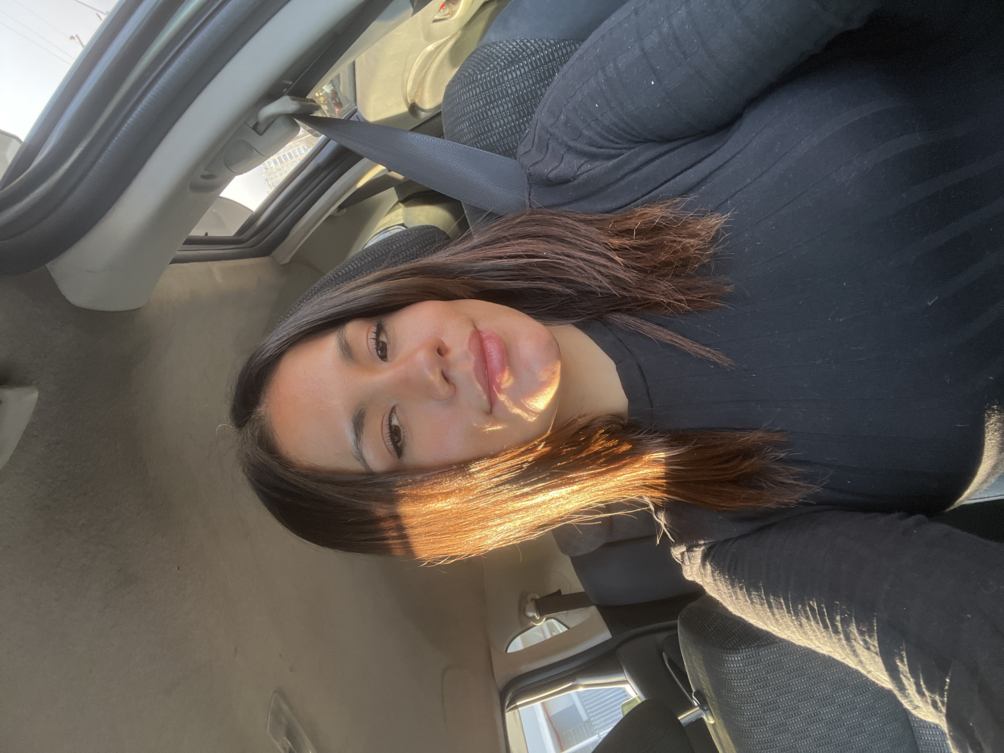 A woman is depicted sitting inside a car, likely in the front passenger seat, with a seatbelt visible across her chest. She is looking directly at the camera with a neutral to subtly pleasant expression, her lips appearing glossy. Her long, dark brown hair is styled down, with some strands catching the light. She is wearing a black ribbed long-sleeved top and what appear to be black pants or jeans.
The setting is the interior of a car, featuring gray-black car seats, a light-colored roof lining, and window frames. Bright, warm sunlight streams in from the side, casting a strong golden glow and highlights on the woman's hair, face, and clothing, indicating a sunny daytime scene. Shadows are also prominently cast across parts of her and the car's interior. Through the car windows, hints of buildings or an outdoor street scene can be vaguely discerned. No visible text is present in the image.