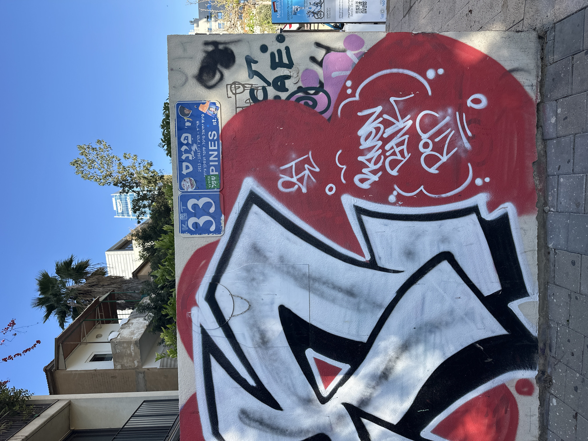 An upright, daytime shot captures a vibrant urban scene in Tel-Aviv, Israel, dominated by a wall extensively covered in graffiti and street signs. The clear blue sky fills the upper left portion of the image, with a green tree and a white residential building visible below it, along with a distant building in the background.
The focal point is the wall, which features layers of street art. A prominent, large red heart shape with white, stylized graffiti and scribbles ("PARDON" and other illegible text) covers a significant part of the wall. Below this, a bold, angular graffiti piece in black and white with red accents takes up the lower section. Above the red heart, there are more tags in black, gray, and pink, including what appears to be "SAVE" and "love".
Two blue street signs are affixed to the wall. The upper sign clearly displays "PINES st." in English and "פינס" (Pines) in Hebrew, along with the biographical details of "פינס יחיאל מיכל. סופר ועסקן ציוני" (Pines Yechiel Michel. Writer and Zionist activist) and the dates "תר"ד - תרע"ג / 1843-1913". It also has a small sticker featuring an anime-style character. Below this, another blue sign shows the number "33," indicating a building number. Parts of what might be another tag or a different sign, "כדורגל" (football/soccer) and "שפל" (low/shallow), are also visible near the Pines street sign.
In the upper right, partially obscured posters feature text like "Metrofu," "No Traffic. No Problem.," "Rent In The App," and Hebrew phrases such as "הקלאסים" (The Classics) and "להיות חברים שלנו" (to be our friends). The bright sunlight casts distinct shadows, indicating a sunny day. The right side of the image shows a transition to a gray, block-paved surface.