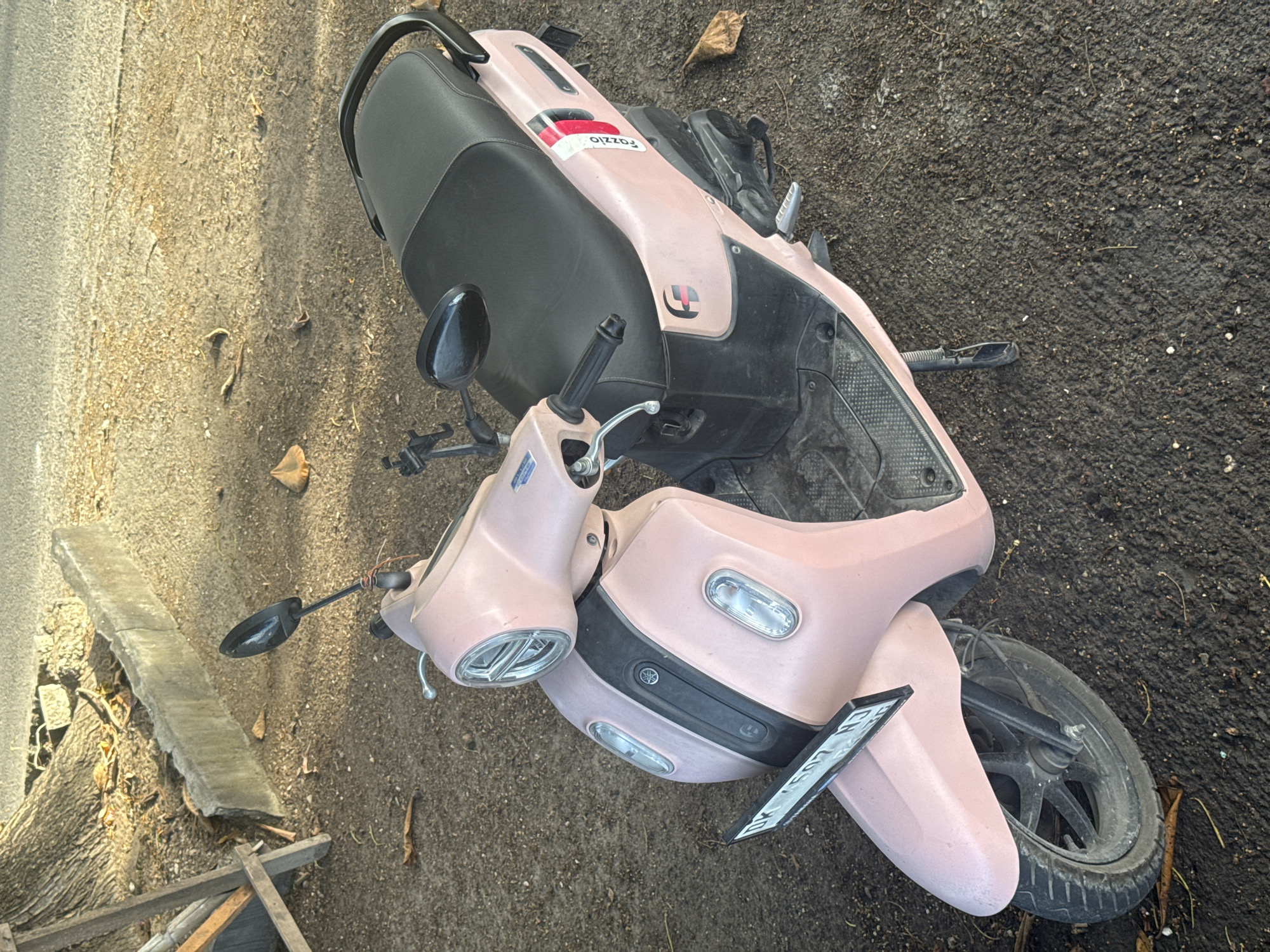 An overhead shot shows a light pink Yamaha Fazzio scooter parked on a dirt and gravel surface next to a paved road in Nusa Penida, Indonesia. The scooter's black seat and handle grips are visible, as is a license plate. A tree trunk and curb are to the left of the scooter, and dry leaves are scattered on the ground. The scene is bathed in sunlight, casting shadows.