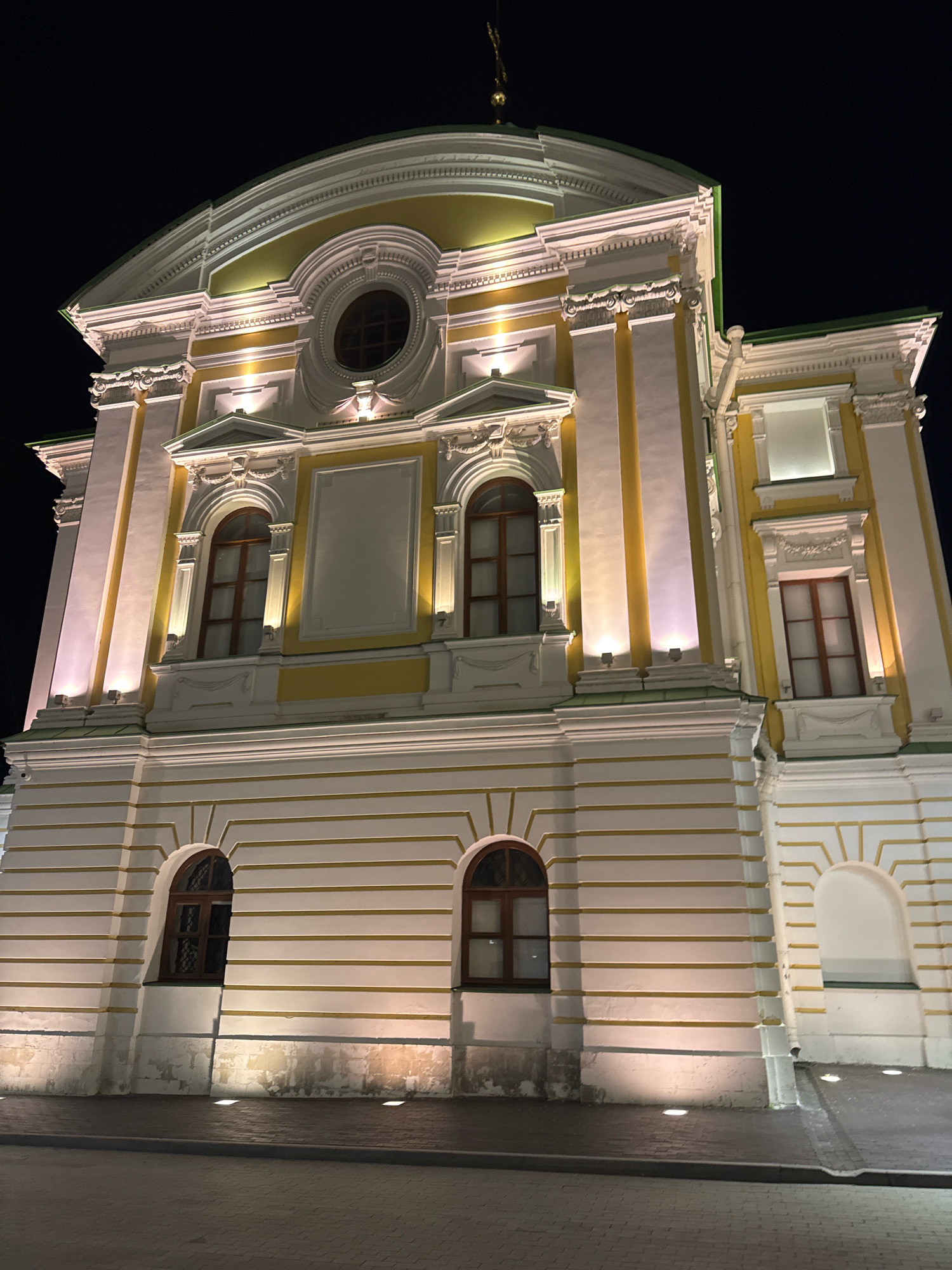 The image shows the illuminated facade of a classical building at night, located in Tver, Russia. The building is white with yellow accents and features ornate architectural details, including columns, decorative molding, and arched windows. Several windows are visible, some with dark wooden frames. The building is lit by warm-toned lights strategically placed to highlight its features. The foreground shows a paved walkway or street with some embedded lights. The sky above is dark, indicating nighttime. There are no people or animals visible in the image. The overall impression is one of grandeur and historical significance, with the lighting emphasizing the building's structure and ornamentation against the dark night sky.