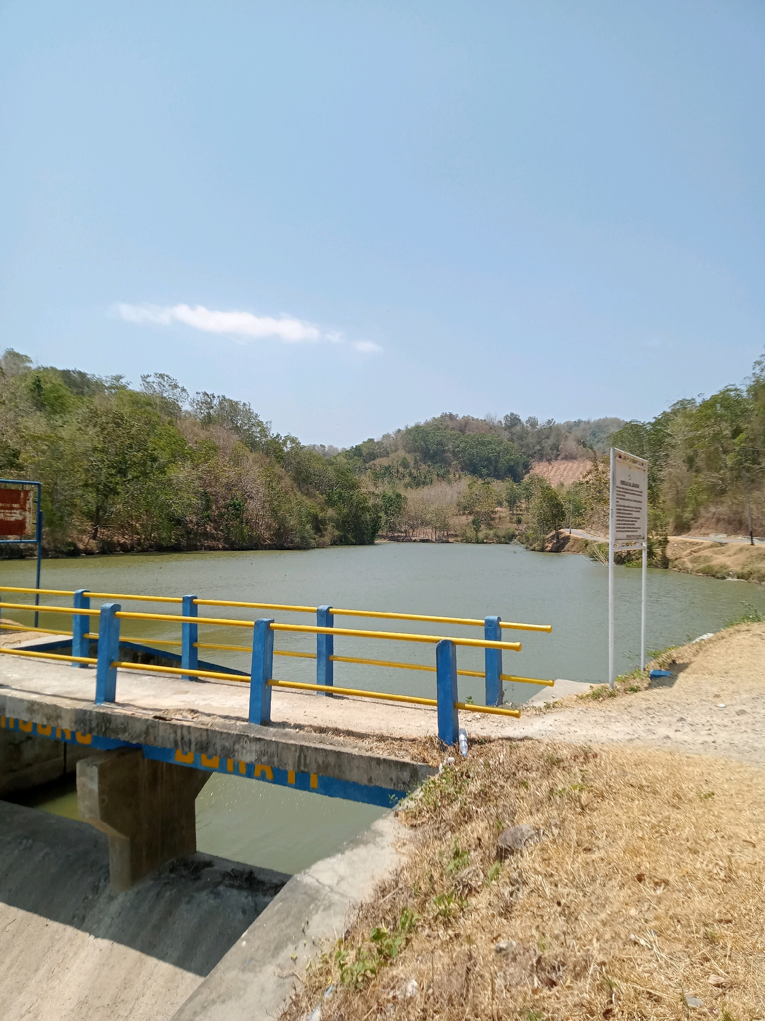 This image depicts a calm reservoir in Luhu, Indonesia, under a clear blue sky. A concrete bridge with blue posts and yellow railings spans part of the reservoir, leading the viewer's eye towards the lush green hills that surround the water. To the right of the reservoir, a white sign with Indonesian text stands near a dirt path. The vegetation is dry and brown in the foreground, suggesting a warm climate. There are no people visible in the image.