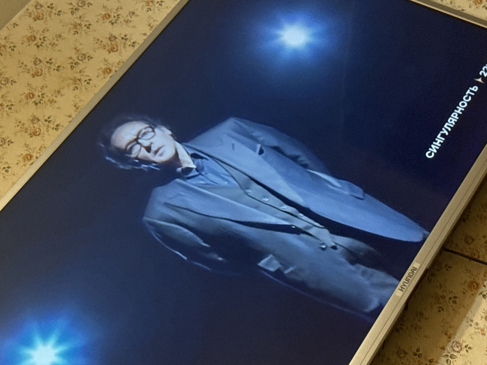 The image shows a television screen displaying a man in glasses and a suit. The screen also displays text in Russian, "СИНГУЛЯРНОСТЬ," and a play button symbol with "22" next to it, suggesting the program is paused at 22 minutes. The lower right corner of the television screen has "HYUNDAI" printed on it. The TV is partially on a surface covered with a floral patterned material. The environment of Mytishchi, Russia is not discernable from the image, therefore no determination can be made on the location.