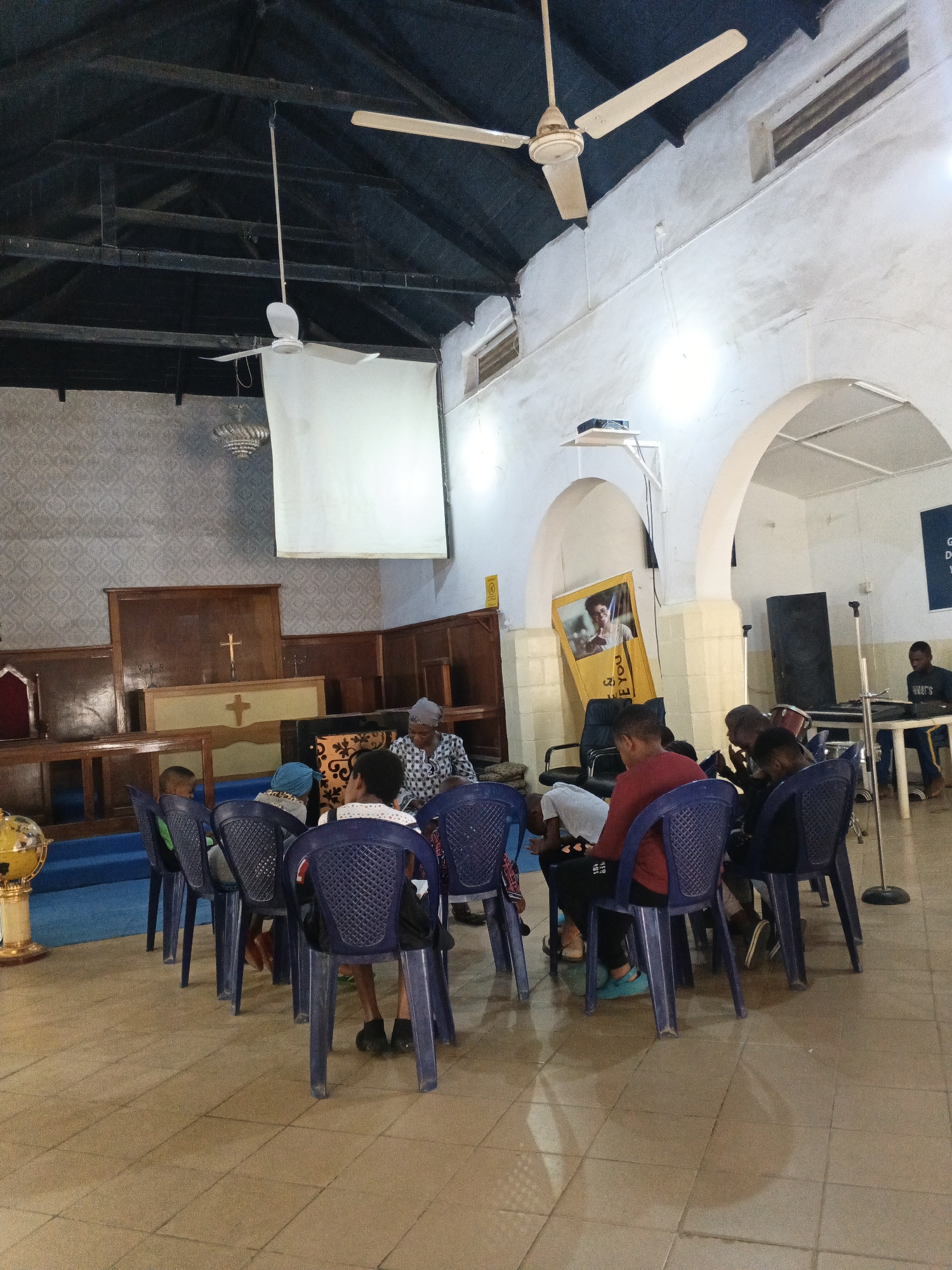 **Scene:** Interior of a building, possibly a church or community center, located in Unguwar Sarki, Nigeria.
**People:** A group of people, including children and adults, are seated in a circle of blue plastic chairs. One person, possibly an older woman, appears to be leading or addressing the group. A single person is sitting at a table in the background.
**Objects:** The room features a wooden altar with a cross, a projector screen hanging from the ceiling, a sound system (speaker and microphone stand), a banner, and a globe. There are ceiling fans visible.
**Actions:** The people are gathered in a circle, likely for a discussion or meeting. The person in the background appears to be working at a table, possibly operating a keyboard.
**Environment:** The building has tiled flooring and white walls with arched openings. The ceiling is dark and exposed, possibly constructed of wood.