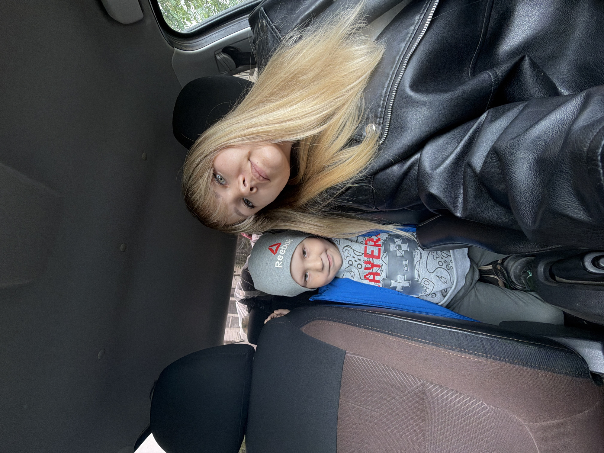 An overhead, sideways-oriented selfie taken inside a car in Chaikovsky, Russia, features a blonde woman and a young boy. The woman, with long blonde hair and light-colored eyes, is visible in the upper part of the image, wearing a black leather jacket and smiling subtly. She appears to be lying down due to the camera's rotation. Below her, a young boy with light hair is also smiling towards the camera. He wears a grey beanie with the "Reebok" logo and a red delta symbol, a grey long-sleeved t-shirt with stylized letters forming what appears to be "PLAYER" amidst a pattern of gaming controller shapes, and a blue jacket.
The car's interior is visible, including a dark grey headliner and car seats upholstered in black and brown patterned fabric. Green foliage can be seen outside the car window, indicating an outdoor, daytime setting, likely with an overcast sky given the soft, diffused light. Both subjects display calm, pleasant expressions, suggesting a relaxed and happy moment.