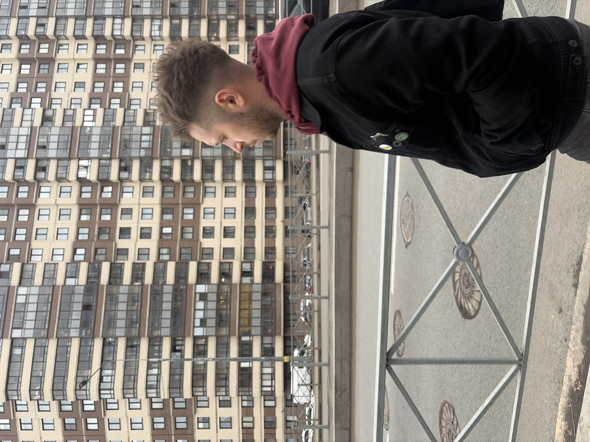 A man in a black jacket and maroon hoodie stands on a sidewalk in Saint Petersburg, Russia, beside a gray metal safety fence. The sidewalk is made of concrete and has metal manhole covers with a seashell-like pattern. A multistory residential building with numerous windows is in the background, and cars are parked on the street below.