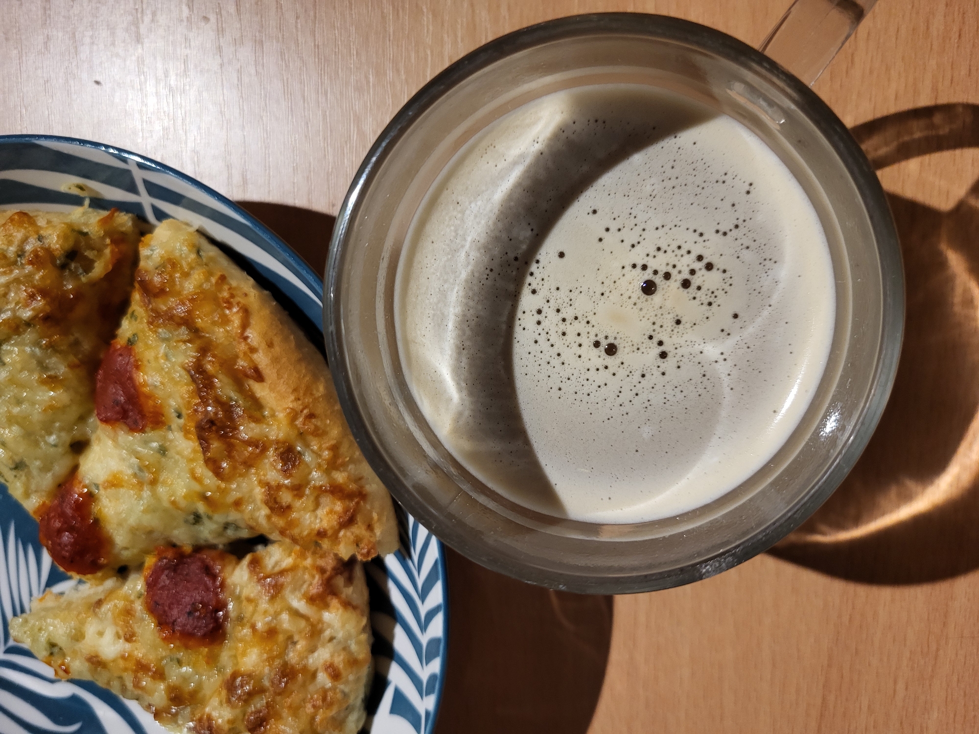A close-up, top-down view shows slices of pizza in a blue and white patterned bowl next to a clear glass mug filled with coffee. The pizza slices are topped with melted cheese and red sauce. The coffee has a layer of foam with small bubbles. The objects are placed on a light brown wooden surface. The scene is lit from above, casting shadows on the table. The location is Unknown, Unknown.