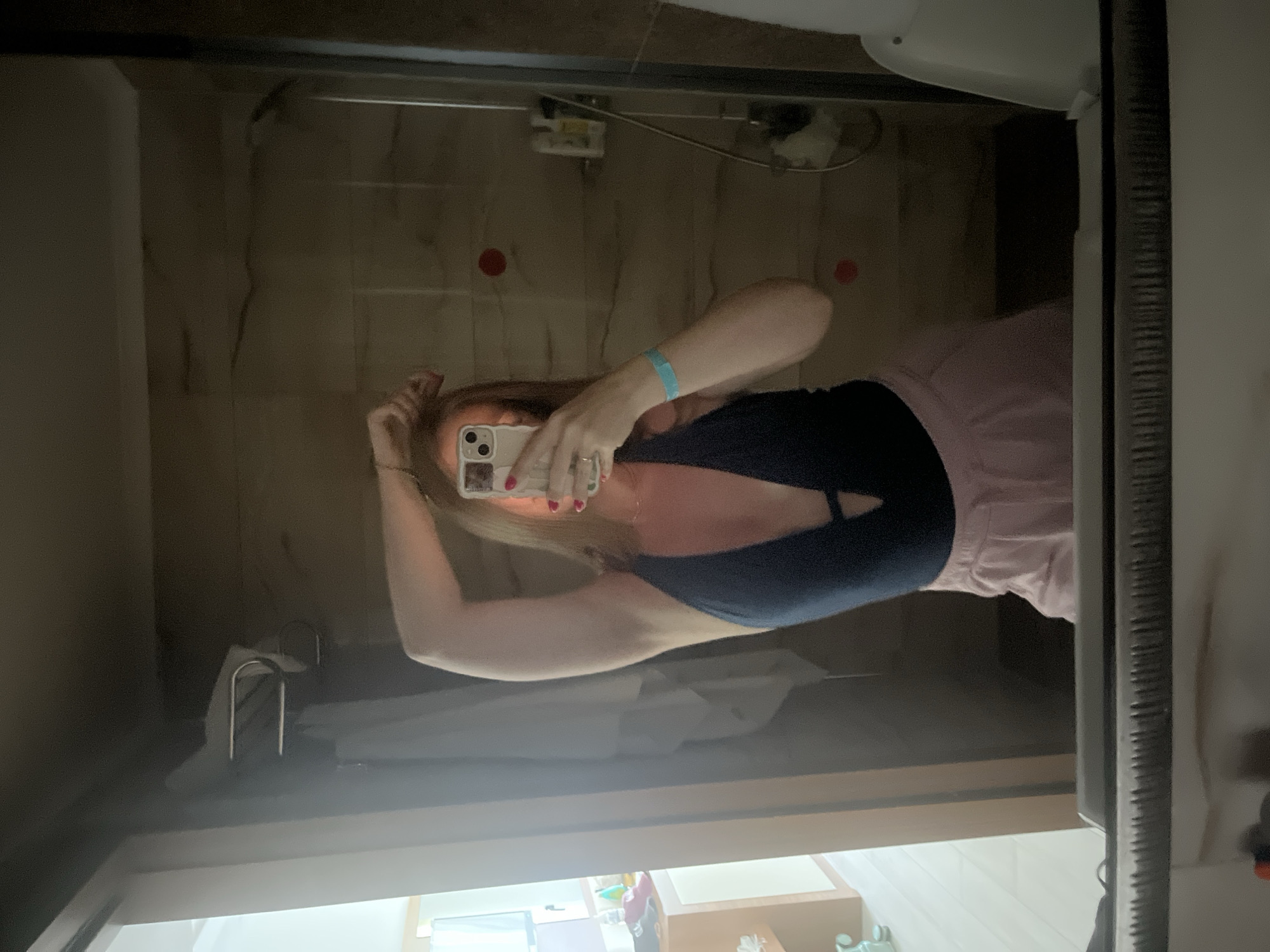 A person is taking a mirror selfie in a bathroom. The person is wearing a dark blue one-piece swimsuit with a deep V-neck and light pink shorts. They have long, light brown hair and are holding a white smartphone in their right hand, angled to capture the reflection. A light blue wristband is visible on their left wrist. The bathroom appears to be in a hotel room, with tiled walls in shades of brown and beige, a glass shower door, and a towel rack with a white towel. In the background, a white toilet and a bidet are partially visible. To the left of the frame, a dresser or vanity with some items on it can be seen, along with a wall-mounted television. The floor is light-colored, possibly laminate or tile. The overall impression is a casual, personal moment captured in a private space.