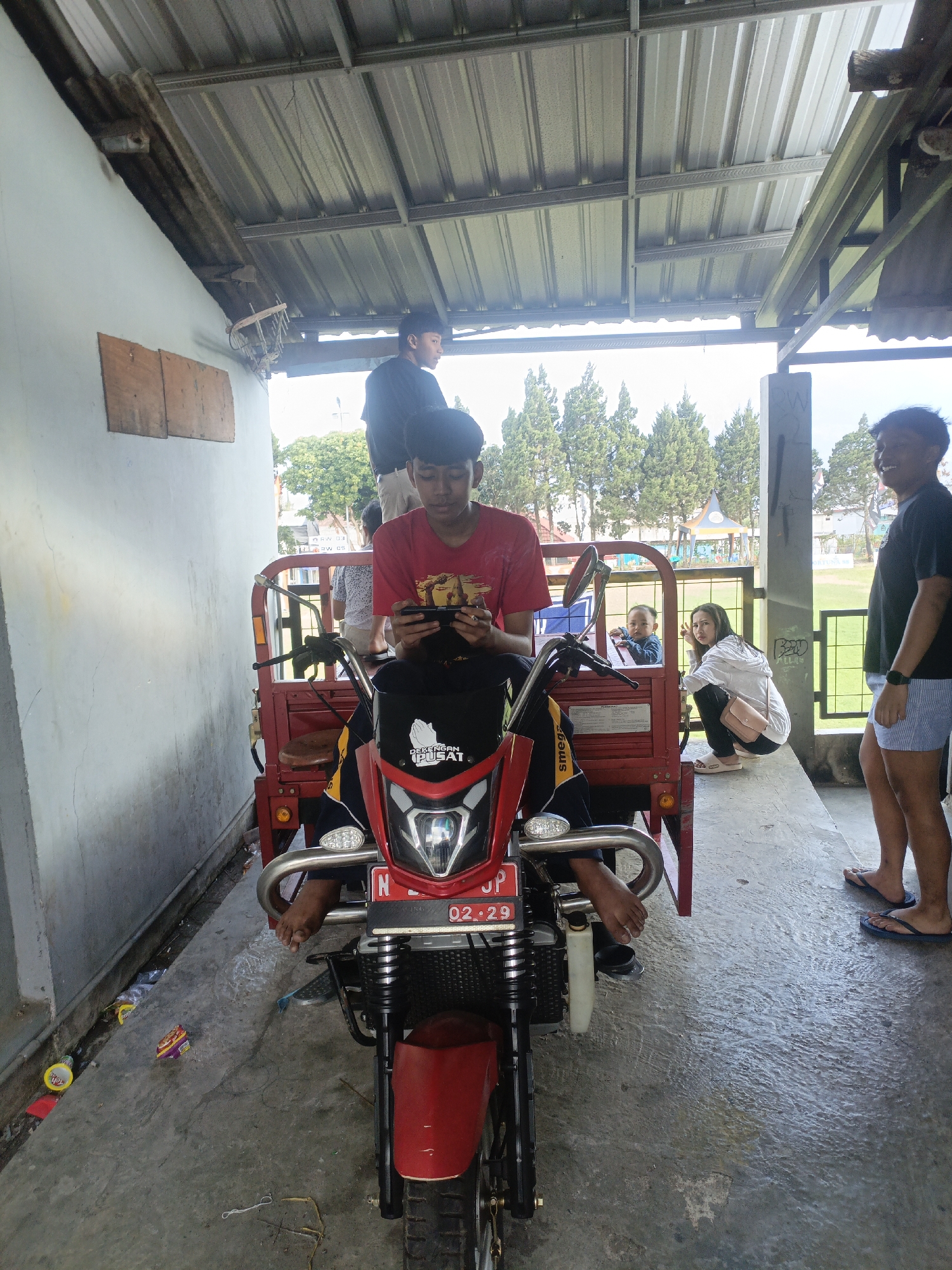 In Batu, Indonesia, an outdoor scene unfolds under a corrugated metal roof supported by metal beams. A red motorized three-wheeled vehicle, commonly known as a "becak motor," is the central focus. A young male, wearing a red t-shirt and black shorts, is seated on the driver's portion of the becak motor, engrossed in playing a mobile game. His legs are visible, bare and resting on the vehicle's frame. To his left, another young male in a black t-shirt stands partially visible.
Behind the driver's seat, a young child wearing a blue t-shirt is seated, looking towards the camera. Adjacent to the child, a young woman in a white hoodie and dark pants is crouched down, facing the child. She has a pink crossbody bag slung over her shoulder. To the right of the becak motor, a young male in a black t-shirt and blue-striped shorts stands with his back to the camera, smiling. The ground is paved with concrete, appearing damp in places, with scattered litter including snack wrappers and plastic bottles.
In the background, a field with lush green trees and a playground structure is visible, suggesting the location is near an open recreational area. The overall atmosphere is casual and social, with people gathered around the becak motor.