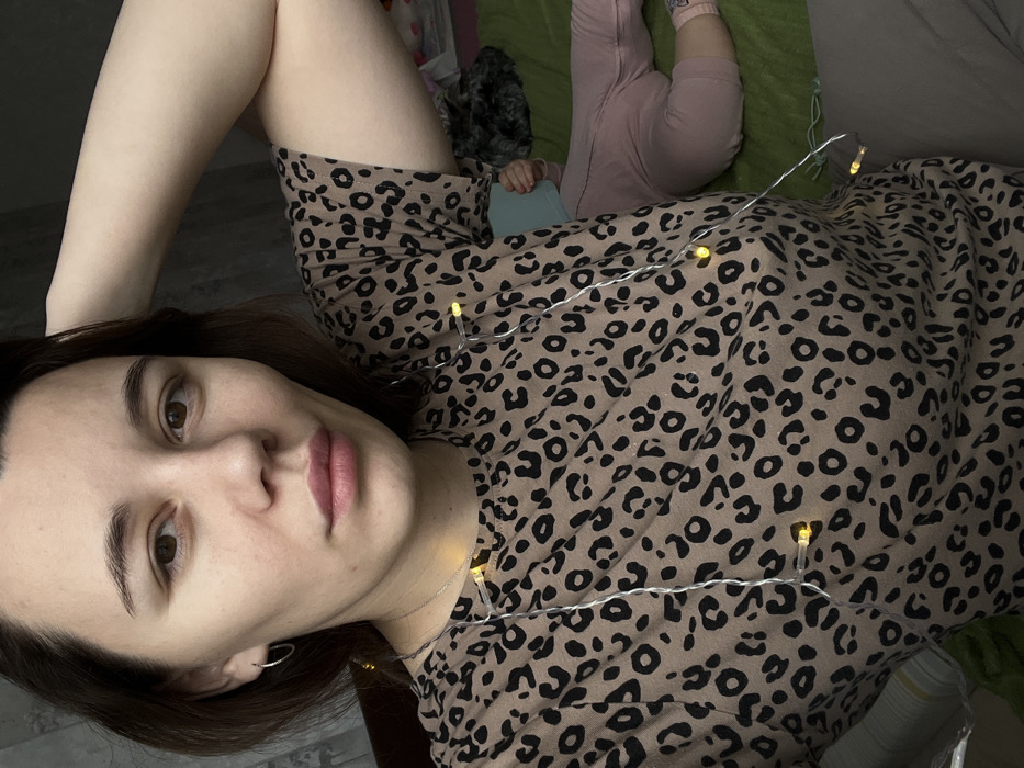 The image depicts a female subject in a casual indoor setting, specified as being within the city of Leninakan, Russia. The woman, possessing dark brown hair and fair complexion, occupies the majority of the frame, looking directly forward at the viewer. She is attired in a beige t-shirt featuring a black leopard print pattern and wears small silver hoop earrings. A string of illuminated warm white LED fairy lights is draped diagonally across her torso, from her left shoulder to her right hip. Her right arm is raised, with her hand positioned near the back of her head. In the lower right background, a child is partially visible, lying supine on a green surface, attired in pink pants and a light-colored sock on the visible left foot. The child's upper body and head are partially obscured by a light blue object. The background immediately behind the woman consists of a dark, textured wall or partition.
