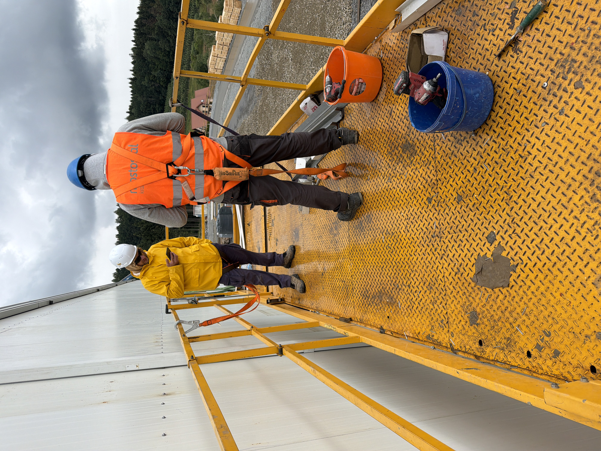Two construction workers are on a yellow elevated platform. The worker on the left, wearing a yellow raincoat and white hard hat, is looking at their phone. The worker on the right, wearing a blue hard hat and an orange safety vest over a gray hoodie, is facing away from the camera. They are wearing safety harnesses. In the foreground, on the yellow platform, there are two buckets, one orange and one blue, containing tools and what appear to be fasteners. In the background, there is a white building, trees, and stacks of lumber. The sky is overcast. The scene is in the city of Stróża, Poland, a location known for its industrial and construction activities.