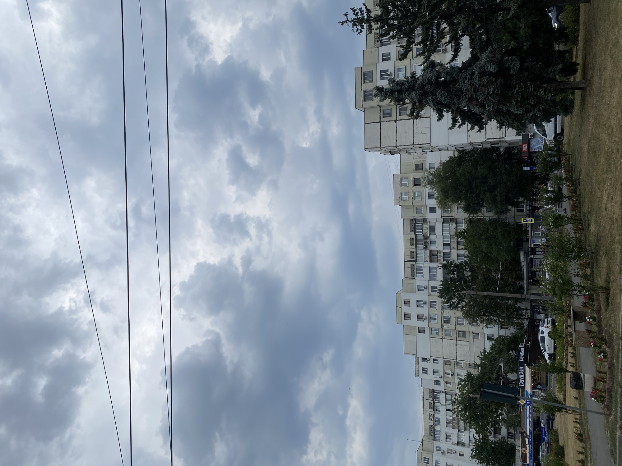 The image depicts a cloudy sky over a long, multi-story apartment building in Chișinău, Moldova. The building appears to be constructed of concrete panels and has many windows. In front of the building, there is a street with traffic, including cars and a bus. Several trees and some green spaces with flower beds are visible in the foreground. Signs for "DavidDan" and "RODOG" are discernible on the storefronts at the base of the building. The overall scene suggests an urban environment under overcast weather conditions.
