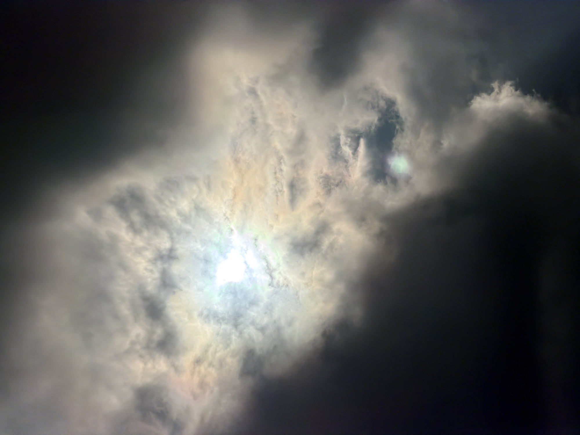 The image depicts a dramatic sky with dark, heavy clouds obscuring a bright, diffused light source, likely the sun, which appears as a pale, luminous disc. The clouds exhibit varying shades of gray, from deep charcoal to lighter, wispy tones, creating a sense of depth and turbulence. Patches of iridescent color, possibly an artifact of light refraction or atmospheric phenomena, are visible within the clouds. The composition is abstract, focusing solely on the celestial elements. The location is identified as Garmisch-Partenkirchen, Germany, suggesting this is a view of the sky above that Bavarian town, likely during a period of inclement weather. There are no people, subjects, objects, or actions visible; the scene is entirely environmental.