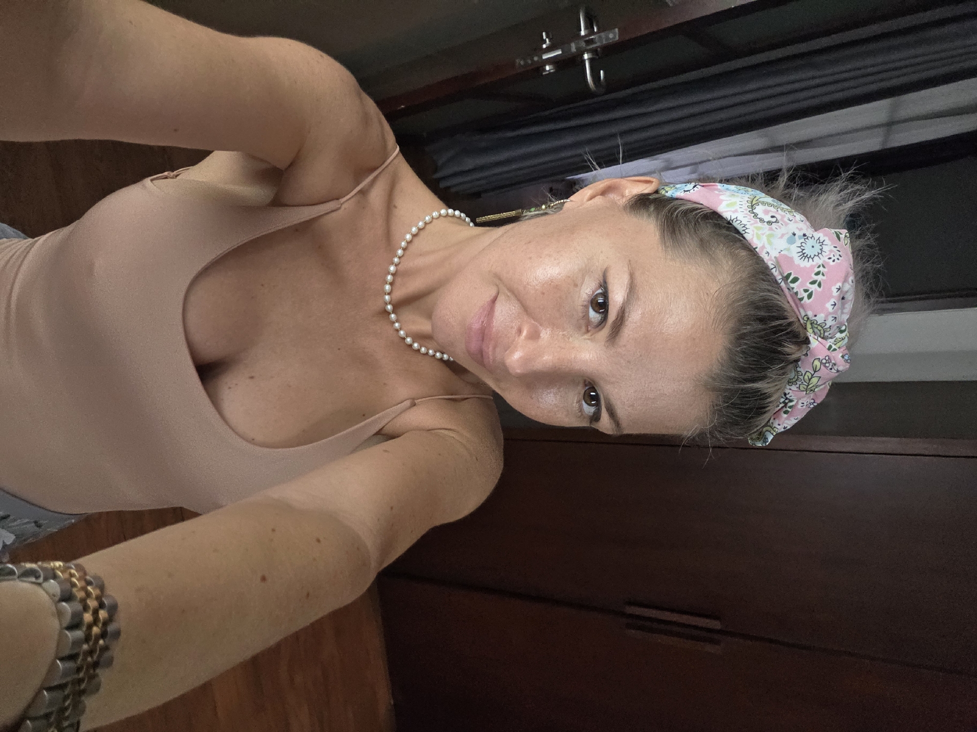 The image shows a woman taking a selfie. She is wearing a pink and white floral headband, a pearl necklace, and a beige spaghetti strap top. Her hair is pulled back, and some strands are sticking out. She is wearing light makeup, including eyeliner. In the background, there is a dark brown wooden closet and a gray curtain with vertical lines. The floor is made of wooden planks. The image appears to be taken indoors, possibly in a bedroom or dressing area. The location of North Kuta, Indonesia is not visible in the image.