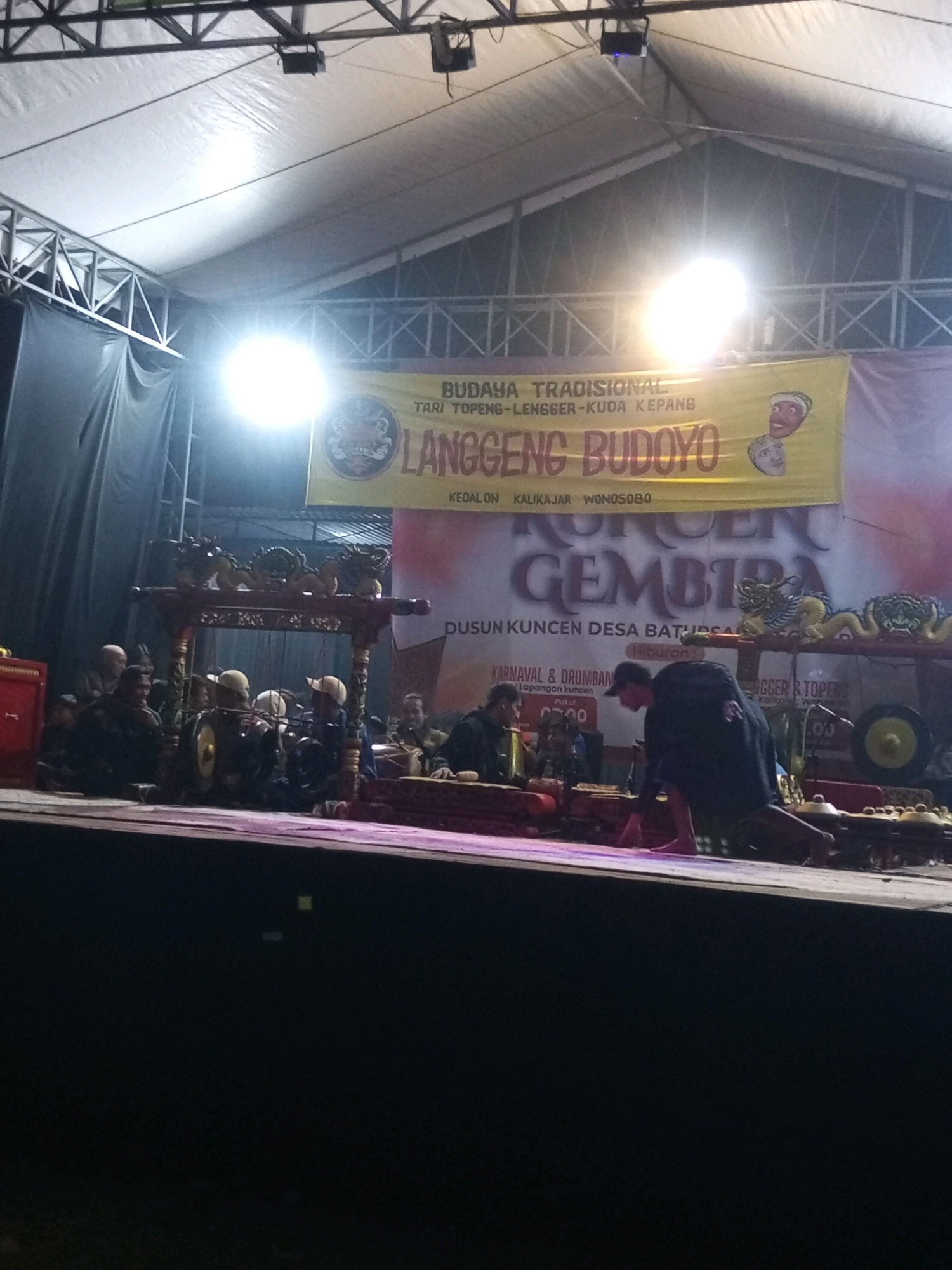 The image depicts a nighttime outdoor performance on a raised stage under a temporary tent structure. A yellow banner with Indonesian text, possibly announcing the event "LANGGENG BUDOYO," is prominently displayed behind the performers. The banner features two cartoonish faces and some decorative elements, including what appears to be a dragon.
Several individuals are visible on the stage. In the foreground, a person wearing a dark outfit is crouched down, facing away from the camera, with bare feet. To the left of this individual, a group of people are seated behind traditional Indonesian musical instruments, likely a gamelan ensemble. These musicians are wearing hats and appear to be playing.
The stage is illuminated by bright spotlights, creating a stark contrast between the lit performance area and the dark surroundings. Various elements of stage equipment, such as curtains and lighting fixtures, are visible. The overall scene suggests a cultural event or festival taking place in Ngadikerso, Indonesia.