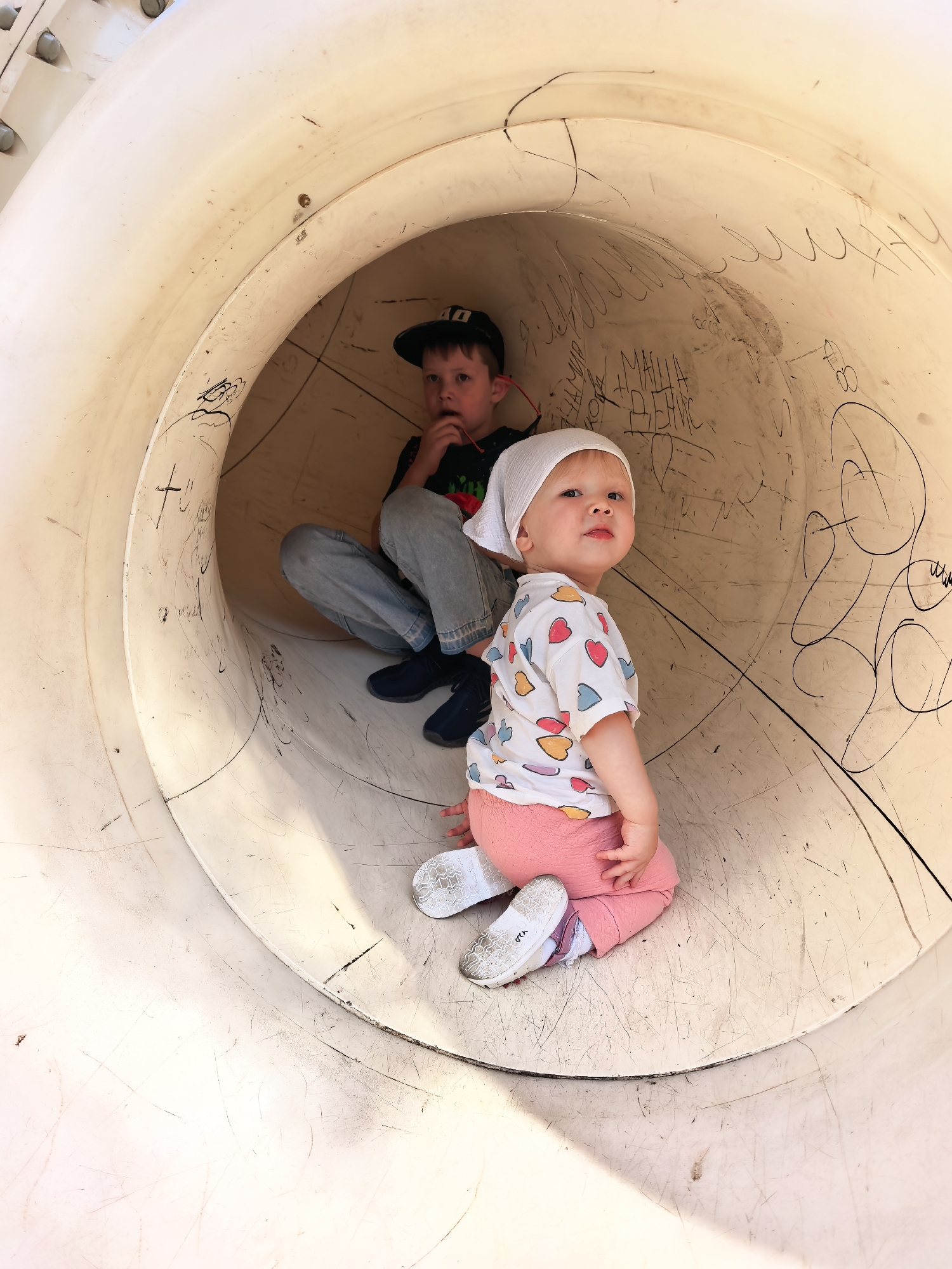 Two children are inside a large, beige, cylindrical tube, possibly a playground slide. The tube has various black markings and scratches on its interior surface.
In the foreground, a young child wearing a white bandana, a white t-shirt with colorful heart designs, and pink pants is sitting facing the right side of the frame, looking towards the camera with an inquisitive expression. The child is wearing white shoes with pink accents.
In the background, an older child wearing a dark baseball cap, a dark t-shirt, and light blue jeans is sitting inside the tube, looking towards the camera and holding something red to their mouth. The child is wearing dark blue shoes.
The lighting suggests an outdoor environment with the sun casting shadows inside the tube. The image appears to have been taken at a playground.
Based on external information, the location is a playground in the city of Sterlitamak, Russia.