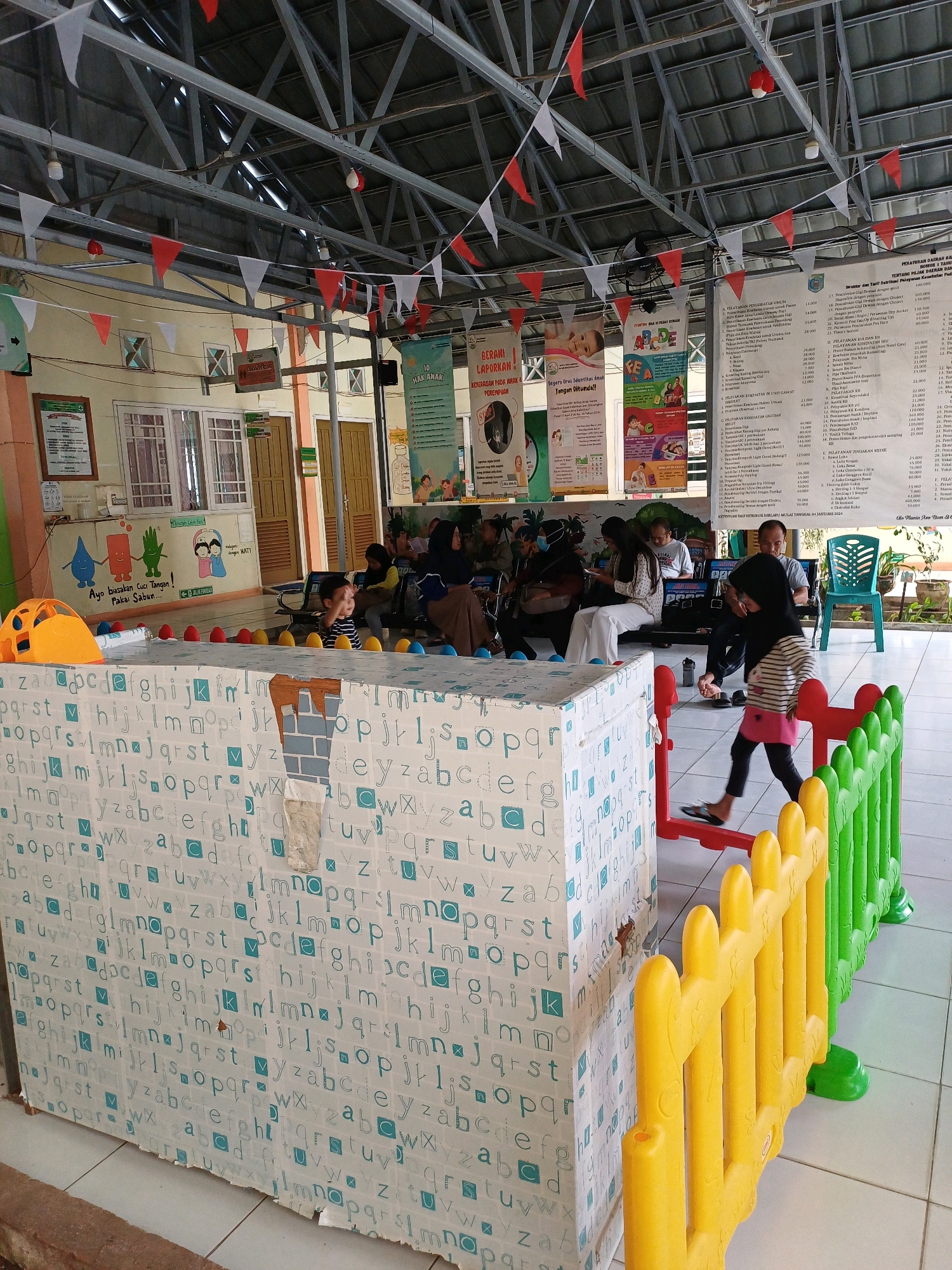 The image depicts an indoor waiting area with several people and children. In the foreground, a low, white playpen is covered in a pattern of blue alphabet letters. Yellow and green plastic safety fences are visible to the right of the playpen, bordering a section of the floor. Several people, including children, are seated on benches or standing in the background, some looking at their phones. A young girl in a hijab and striped shirt is walking past the playpen. In the background, posters and signs are displayed on the wall, and red and white triangular flags are strung across the ceiling. The environment appears to be a public indoor space, possibly a community center or health clinic, given the educational posters and the presence of a play area. The location is identified as Pasir Belengkong, Indonesia.