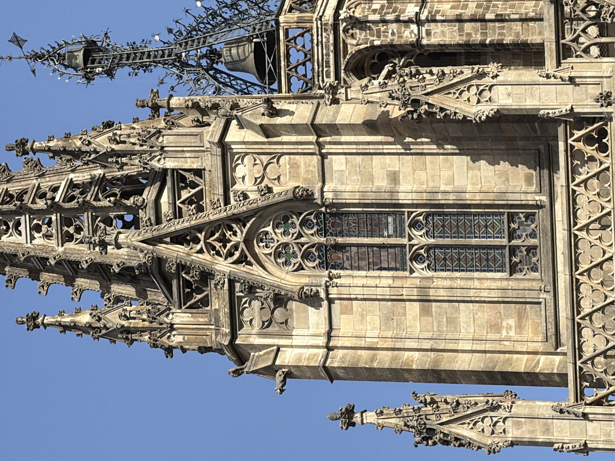 The image captures a close-up view of the intricate gothic architecture of the Barcelona Cathedral, specifically focusing on the upper sections of its spires and facade. The stone structure is bathed in sunlight, casting shadows that highlight the detailed carvings and decorative elements. Visible features include stained-glass windows with geometric patterns, gargoyles perched on ledges, ornate pinnacles, and a bell tower with a weather vane. The sky is a clear, bright blue, providing a stark contrast to the aged stone. The overall impression is one of grandeur and historical significance, showcasing the craftsmanship of medieval architecture against a serene sky.