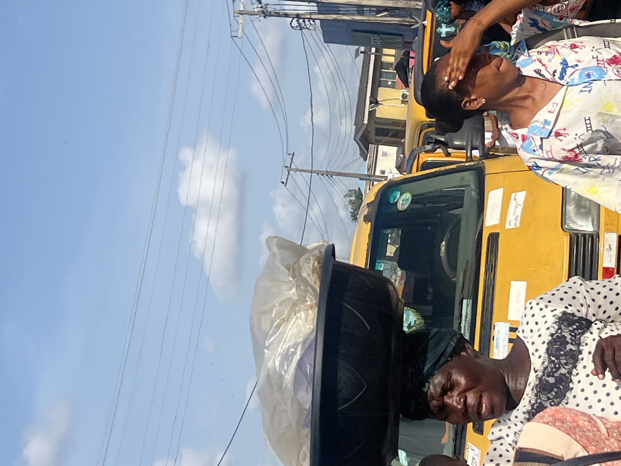 The image depicts a street scene in Egan, Nigeria. A yellow minibus, typical of Nigerian public transport, is visible. Two women are prominent: one wearing a patterned blouse with a hand on her forehead, the other in a polka-dot blouse with a basket on her head containing items covered in plastic. Power lines are visible against a blue sky with scattered clouds.