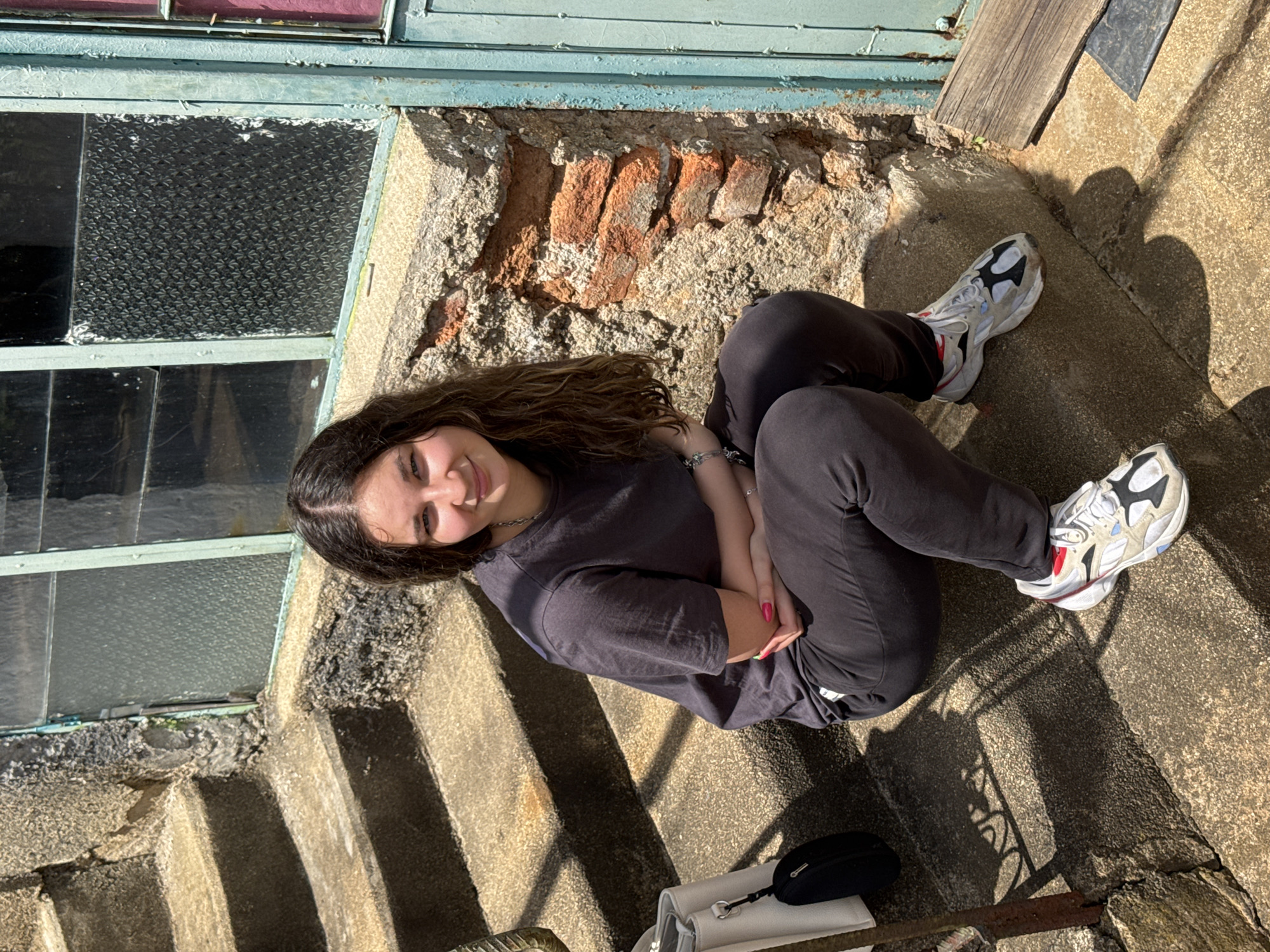 A young woman with long, dark, wavy hair and a light complexion is sitting on concrete stairs in front of a dilapidated building. She is wearing a dark grey t-shirt and dark grey pants, with white, black, and red athletic shoes. Her arms are crossed in front of her chest, and her hands are clasped. The building behind her has a textured, rough-hewn stone facade and a window with diamond-patterned glass. The stairs are also made of concrete and show signs of age and wear. A white purse and a black pouch are placed on the stairs to her left. The scene is outdoors, with strong sunlight casting shadows on the stairs. The overall environment suggests a rural or semi-urban setting in Prolom, Bulgaria, possibly an older or abandoned structure.