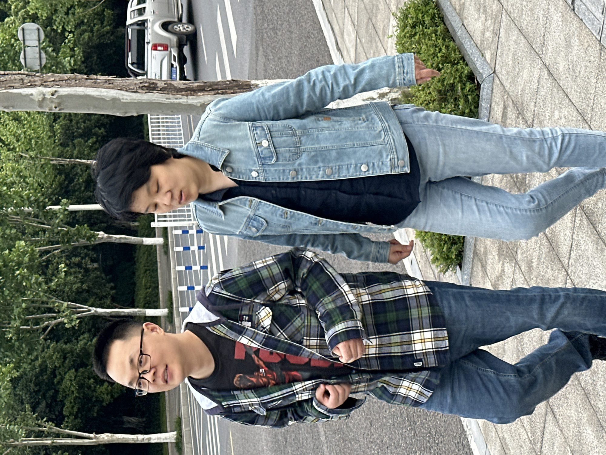 Two individuals stand on a sidewalk in Jiangjin, China. One wears glasses, a black t-shirt with a red graphic, a plaid shirt, and jeans. The other person has short black hair and is wearing a denim jacket, a dark shirt, and jeans. A street with a parked silver pickup truck is visible in the background, along with trees and green bushes. The sidewalk is made of light gray stone blocks.