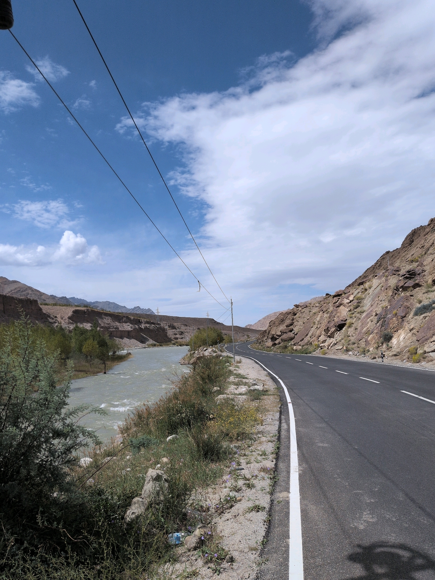 This is a landscape photograph taken in Upshi, India. The scene features a paved road curving to the right, bordered by a rocky, mountainous terrain on the right and a river on the left. The river appears to be flowing swiftly, with visible ripples on its surface. Along the riverbank, there is vegetation, including trees and bushes. In the background, more mountains stretch across the horizon, and the sky is partly cloudy with a bright blue expanse. Power lines are visible in the upper portion of the image, running diagonally across the frame. The road has white lane markings. The overall environment suggests a remote, possibly arid region.