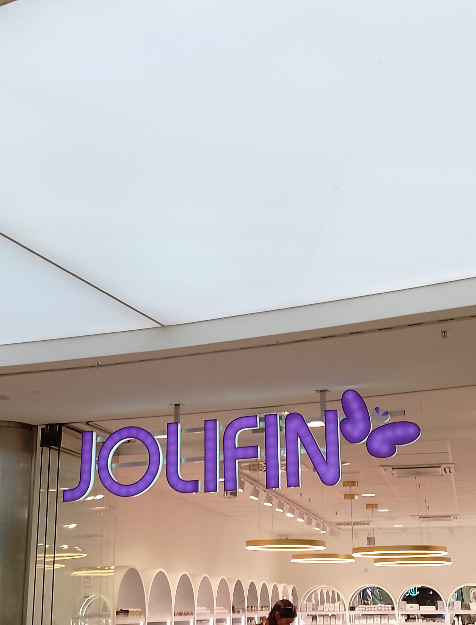 The image shows the exterior of a store with a prominent purple sign that reads "JOLIFIN". The sign is illuminated and features stylized butterflies. Below the sign, the store's interior is visible through large glass windows, revealing a brightly lit space with arched shelves and several hanging circular pendant lights. A woman is partially visible in the lower right corner, looking down. The overall impression is of a modern retail environment, likely a beauty or cosmetics store. The lighting suggests it is daytime, and the indoor setting implies a controlled environment, so weather is not a factor. There are no explicit location cues beyond it being a commercial establishment. The text "JOLIFIN" is clearly visible on the signage.