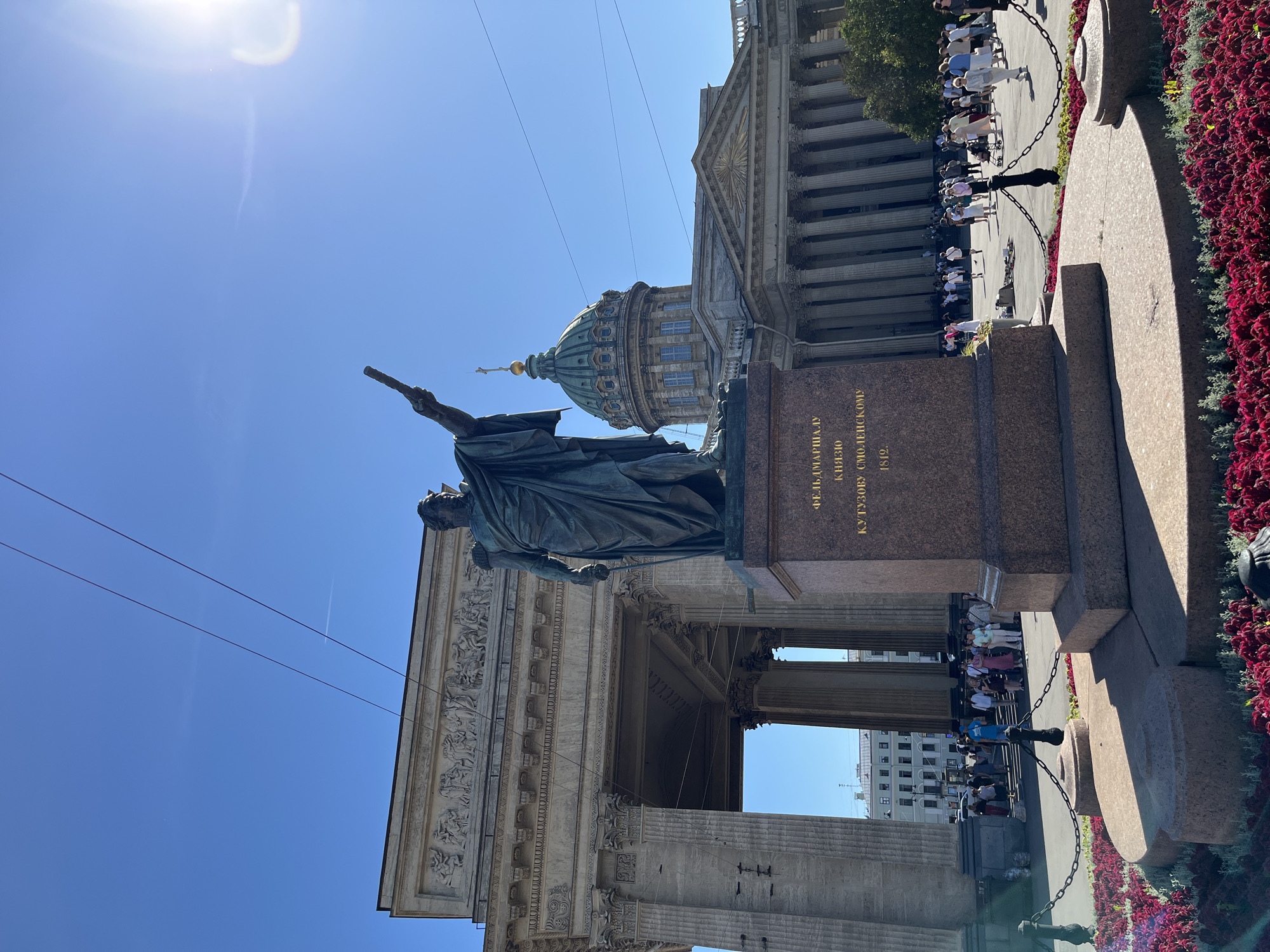 The image depicts a statue of Mikhail Kutuzov in front of the Kazan Cathedral in Saint Petersburg, Russia. The statue, made of bronze, stands on a granite pedestal inscribed with Russian text. The Kazan Cathedral, a prominent landmark in the city, features a grand colonnade and a green dome. The sky is clear and blue, with the sun shining brightly. Several people are visible in the background, walking around the plaza and near the cathedral. The scene is set in a public outdoor space, with red flowers planted in the foreground.