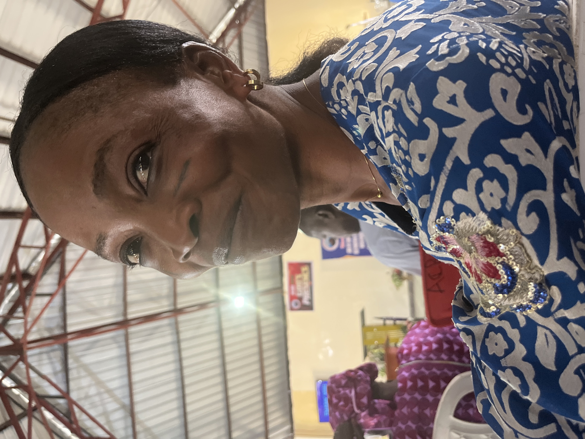 The image shows a woman with dark skin and short black hair. She has gold hoop earrings. She is wearing a blue and white patterned dress with a sequined and embroidered embellishment on the chest. The background suggests an indoor space with a metal roof structure. Other people are visible in the background, seated in chairs. The location is Barakin Akawo, Nigeria.