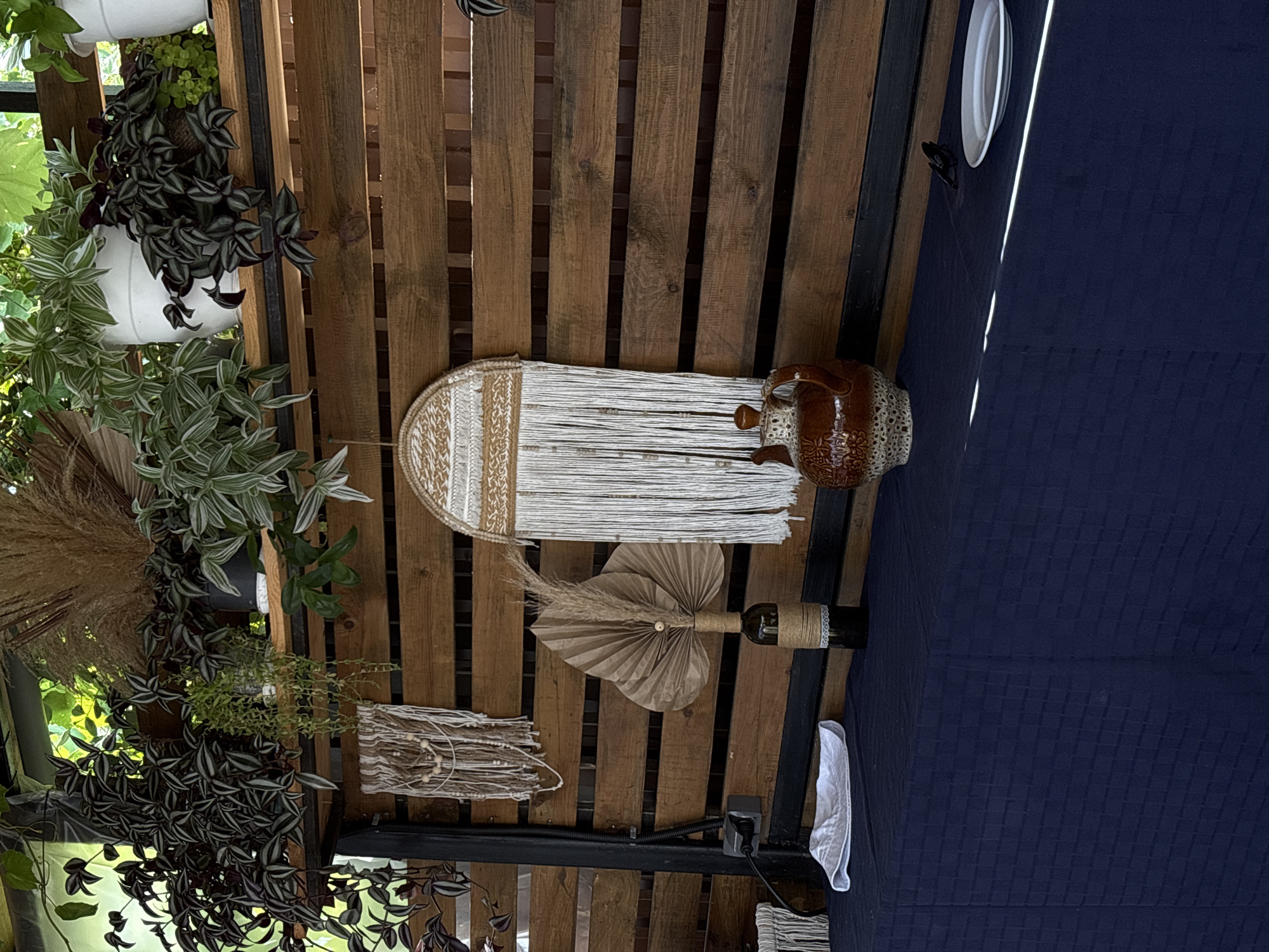 This image captures a rustic and decorative display, likely in an outdoor setting given the natural light and plants. The primary subjects are various decorative items and lush greenery. Among the objects are a macrame wall hanging with white and beige fringe, a rustic brown ceramic jug with intricate patterns, a wine bottle wrapped in twine and adorned with a dried fan-shaped leaf and some dried grass, and another smaller woven decorative piece with beads. Potted plants with variegated leaves, including trailing varieties with dark purple and silver stripes, are strategically placed, adding a vibrant natural element.
The scene appears to be set on a wooden slatted structure, possibly a pergola or a decorative wall, providing a textured backdrop. A dark blue fabric, likely a tablecloth or drape, covers a surface to the right, with a white plate visible on top and a white cloth folded neatly beside it. The lighting suggests it is daytime, with sunlight filtering through, casting some shadows. There are no people visible, nor is any specific activity taking place; the image seems to be a still life composition highlighting the decorative arrangement.
The overall aesthetic is bohemian and natural, with a focus on handcrafted and organic textures. The location context of Gudauta, Abkhazia, evokes a sense of a possibly coastal or garden setting. There is no visible text in the image.