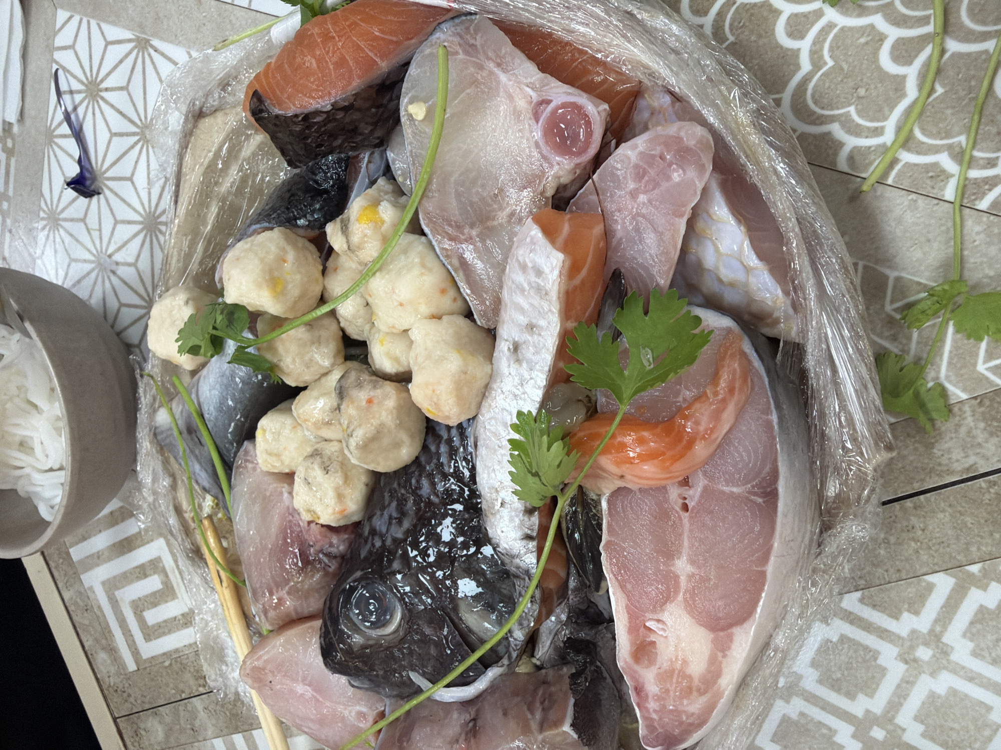 This top-down photograph displays a generous assortment of fresh, raw seafood and fish products, likely prepared for a communal meal such as a hotpot. The collection of ingredients is presented in a clear plastic wrap, possibly for delivery or to maintain freshness, resting on thin wooden skewers or chopsticks.
The prominent subjects are various fish cuts, including several pieces of pink-orange salmon, lighter white fish fillets, and a whole dark-skinned fish head with a visible eye. Mixed in are numerous light-colored fish balls, some speckled with orange or yellow, and a bright orange piece of shrimp. Fresh green sprigs of cilantro or a similar herb are interspersed among the seafood, providing a touch of garnish.
To the left of the main seafood platter, a light grey bowl holds a portion of white, thin noodles, commonly used in hotpot dishes. The setting is a surface adorned with two distinct patterned tiles: one with a white background and an intricate geometric design, and another with a beige tone featuring a softer, curved, floral-like pattern. A small, delicate blue flower rests on the white patterned tile in the upper left.
The image suggests an imminent cooking activity, highlighting the variety and freshness of ingredients. The scene is likely an indoor setting, given the clear, bright lighting. No text is visible in the image. The culinary presentation aligns with dining practices found in Đà Nẵng, Vietnam.