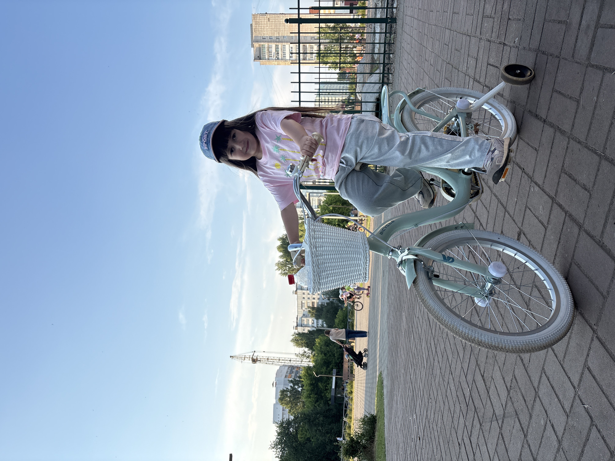 A young girl with long, dark hair wears a pink t-shirt, grey sweatpants, and a blue baseball cap. She sits on a light teal bicycle with a white wicker basket on the handlebars, which contains a clear plastic bottle with a red cap. The bicycle has training wheels. The girl is posed looking at the camera with a slight smile, her hands on the handlebars. The bicycle is on a paved walkway made of grey bricks. In the background, several residential buildings are visible, along with trees, a construction crane, and a few other people and bicycles. The sky is blue with a few scattered clouds. The image was taken in the city of Chaikovsky, Russia.