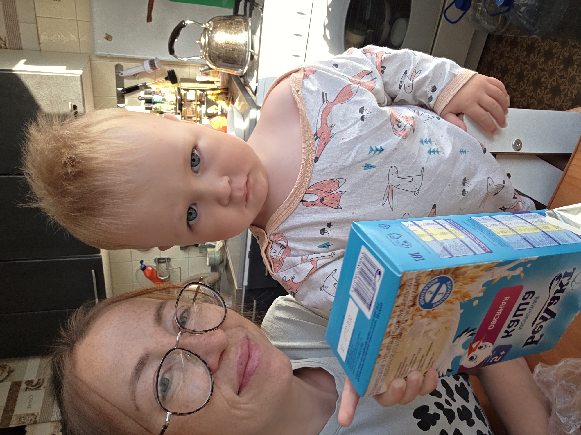 A woman and a child are in a kitchen. The woman has short red hair, fair skin, and round glasses. She is looking at the camera and smiling slightly. She is wearing a white shirt with black spots. The child is sitting in a highchair and looking at the camera. The child has blond hair, blue eyes, and fair skin. The child is wearing a white and pink onesie with fox and bunny patterns. The woman is holding a blue box with Russian writing and an image of a stork on it. In the background, there is a kitchen with a stainless steel kettle, cabinets, and other kitchen items. The image was likely taken in a residential setting. The city of Chaikovsky, Russia, cannot be definitively determined from the image.