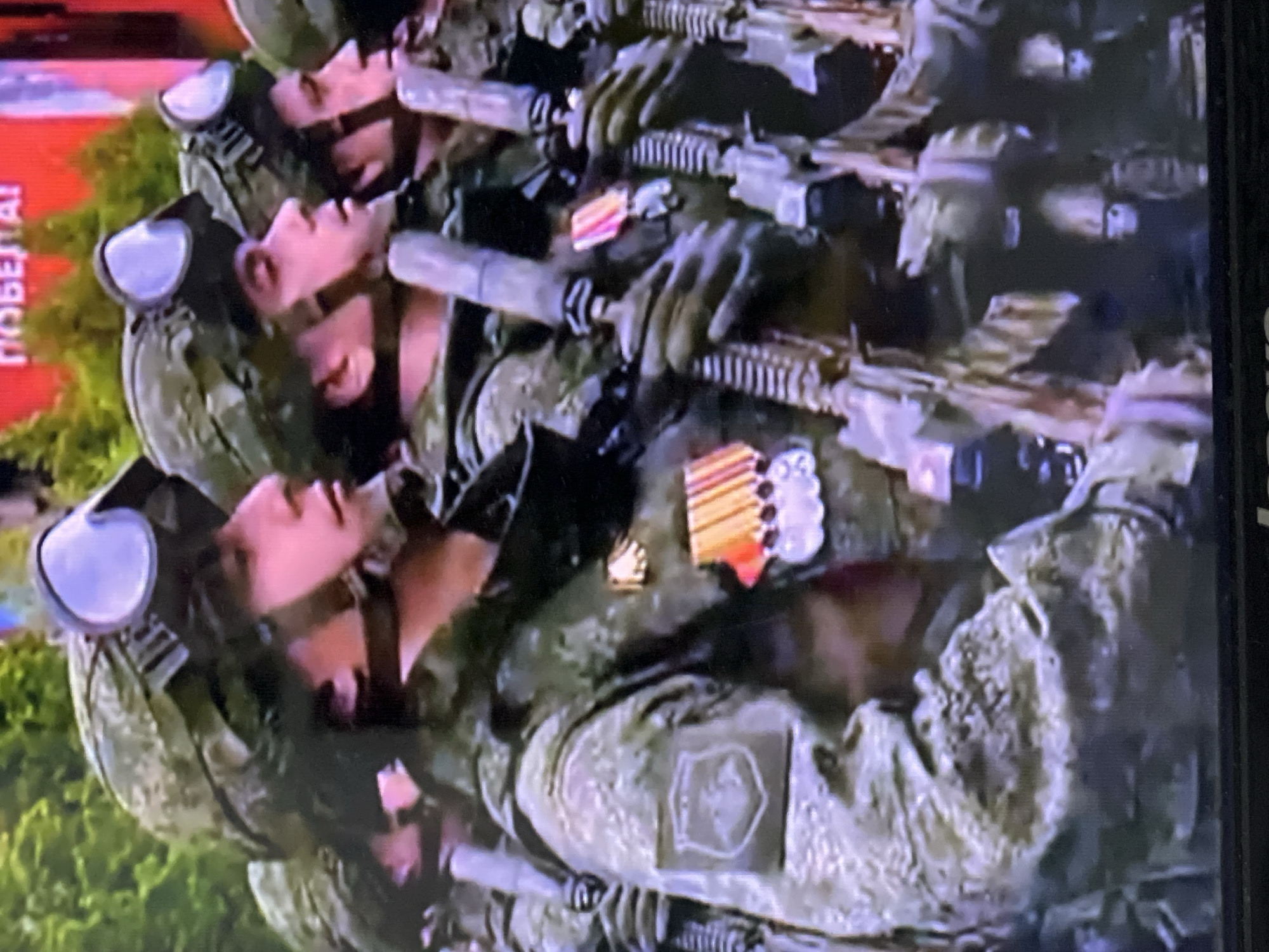 A photograph shows a row of Russian soldiers in camouflage uniforms holding rifles, likely during a military parade or ceremony. The soldiers are wearing helmets and goggles. Some have medals or ribbons on their uniforms. One soldier's uniform has a patch on the arm. The location is not discernible from the image itself. The city of Chaikovsky, Russia, is not evident in the image.