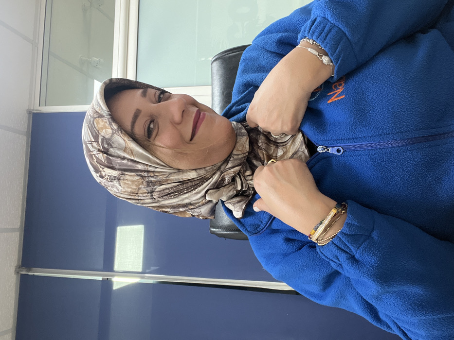 An adult female subject is depicted from the upper chest upward, seated in what appears to be an office environment in Meknes, Morocco. She is wearing a multi-toned patterned hijab, primarily beige and brown, over a royal blue fleece jacket with a front zipper. A partial logo, "Naz...", is visible on the right chest area of the jacket. The subject has dark hair visible beneath the hijab, dark eyes, and dark red lipstick, and is smiling towards the viewer. Her hands are raised, with fingers resting on the lapels of her jacket. On her left wrist, multiple bracelets are visible, including beaded and stone-embellished types. On her right wrist, a silver-toned beaded bracelet with a charm is present, and a gold-toned ring is on her right ring finger. The background features a dark blue vertical panel to the left, and a window or glass partition to the right, reflecting overhead lighting. A black office chair back is partially visible behind the subject. The ceiling consists of white acoustic tiles with integrated lighting fixtures.