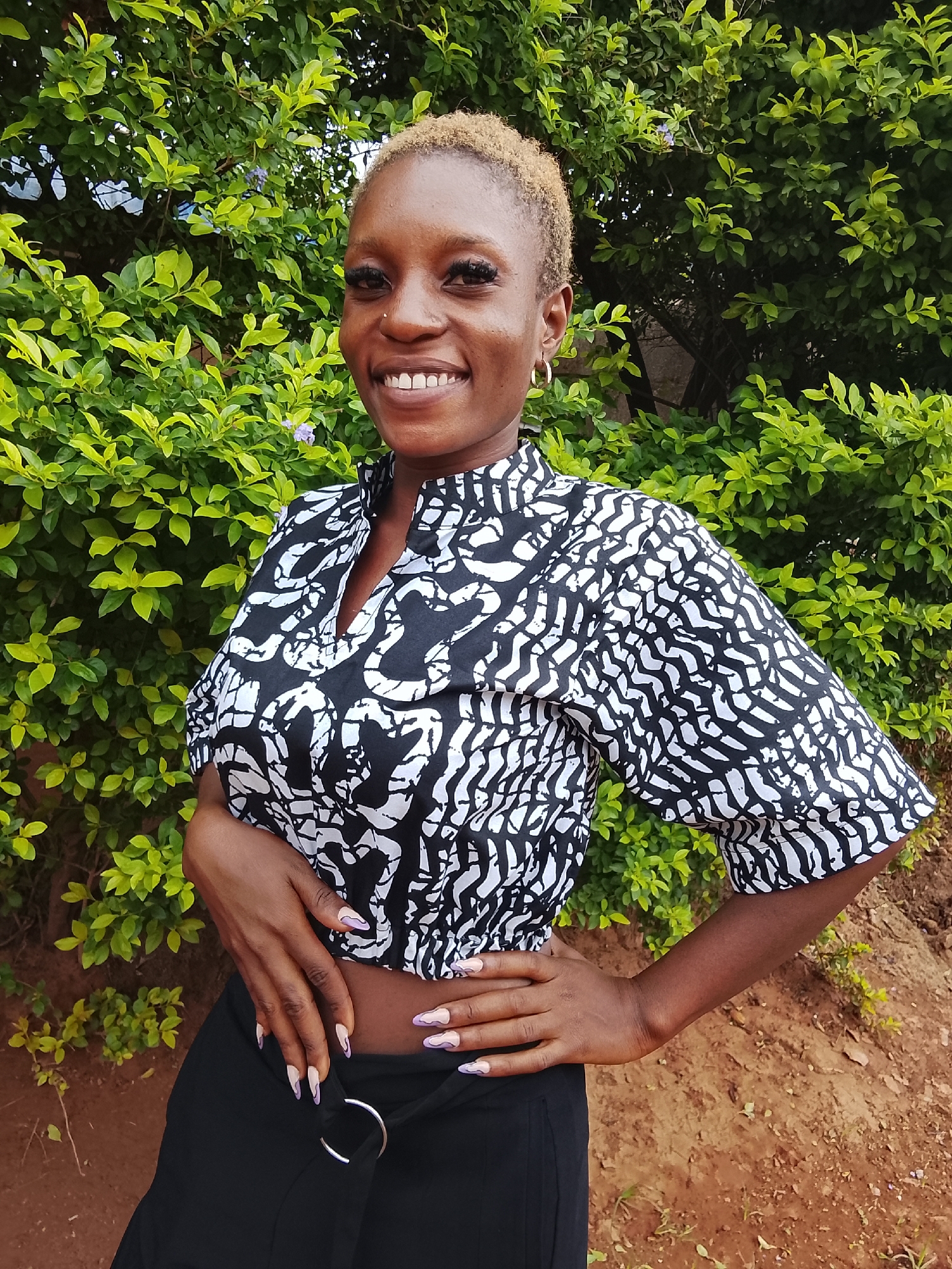 A woman stands outdoors in what appears to be Kuru Karama, Nigeria. She has short, light blonde hair and dark skin. She wears a black and white patterned cropped top and black pants with a silver ring detail at the waist. She is smiling, with her hands on her waist and lower abdomen. Behind her is a lush green bush. The ground appears to be reddish dirt. She has a nose piercing. Her fingernails have a distinctive manicure.