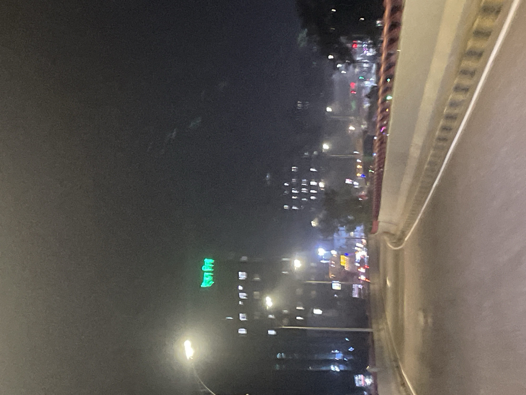 The image captures a nighttime urban scene in Dhaka, Bangladesh, seemingly taken from an elevated position, possibly from a bridge or high building. The right side of the frame shows the edge of a roadway, with visible grid-like patterns on the bridge structure. In the background, illuminated buildings and streetlights punctuate the darkness. A green neon sign stands out on one of the buildings. Other buildings with multiple illuminated windows dot the skyline. A street lamp is visible in the foreground. The general environment appears foggy or hazy, obscuring clarity and depth. The setting suggests a densely populated urban area with active street lighting and infrastructure.