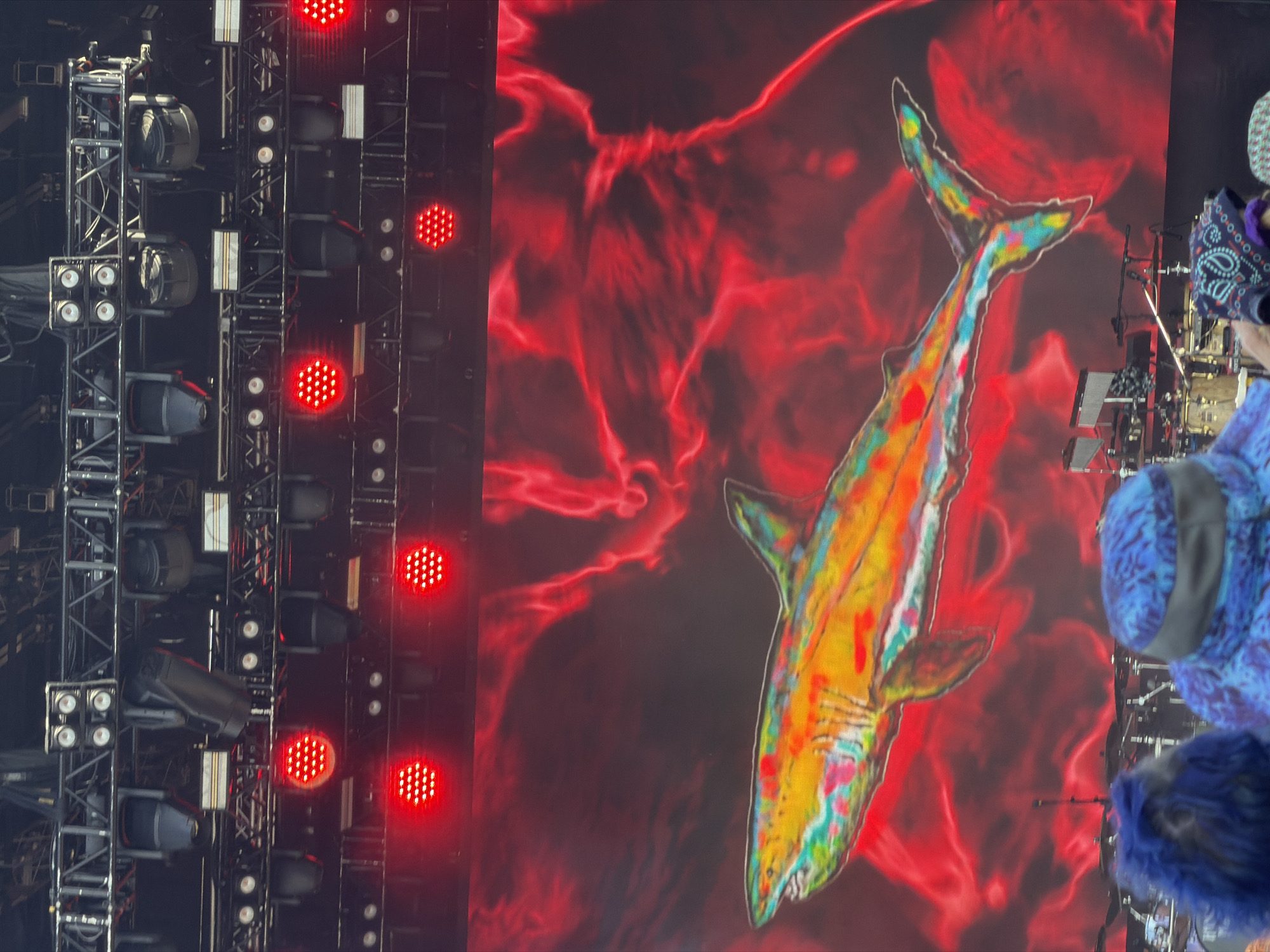 The image captures a vibrant concert scene at the Oceanfront in Virginia Beach, Virginia. A massive LED screen dominates the background, displaying a psychedelic, abstract red pattern with a brightly colored shark illustration. The shark is rendered in a rainbow of hues, including yellow, orange, green, and blue, and appears to be swimming. Above the screen, a complex stage rig with numerous spotlights is visible. Red lights are also illuminating the stage from various positions. In the foreground, the heads and shoulders of concertgoers are seen, some wearing hats and sporting colorful hair. The overall atmosphere suggests an energetic and visually stimulating live music event.
