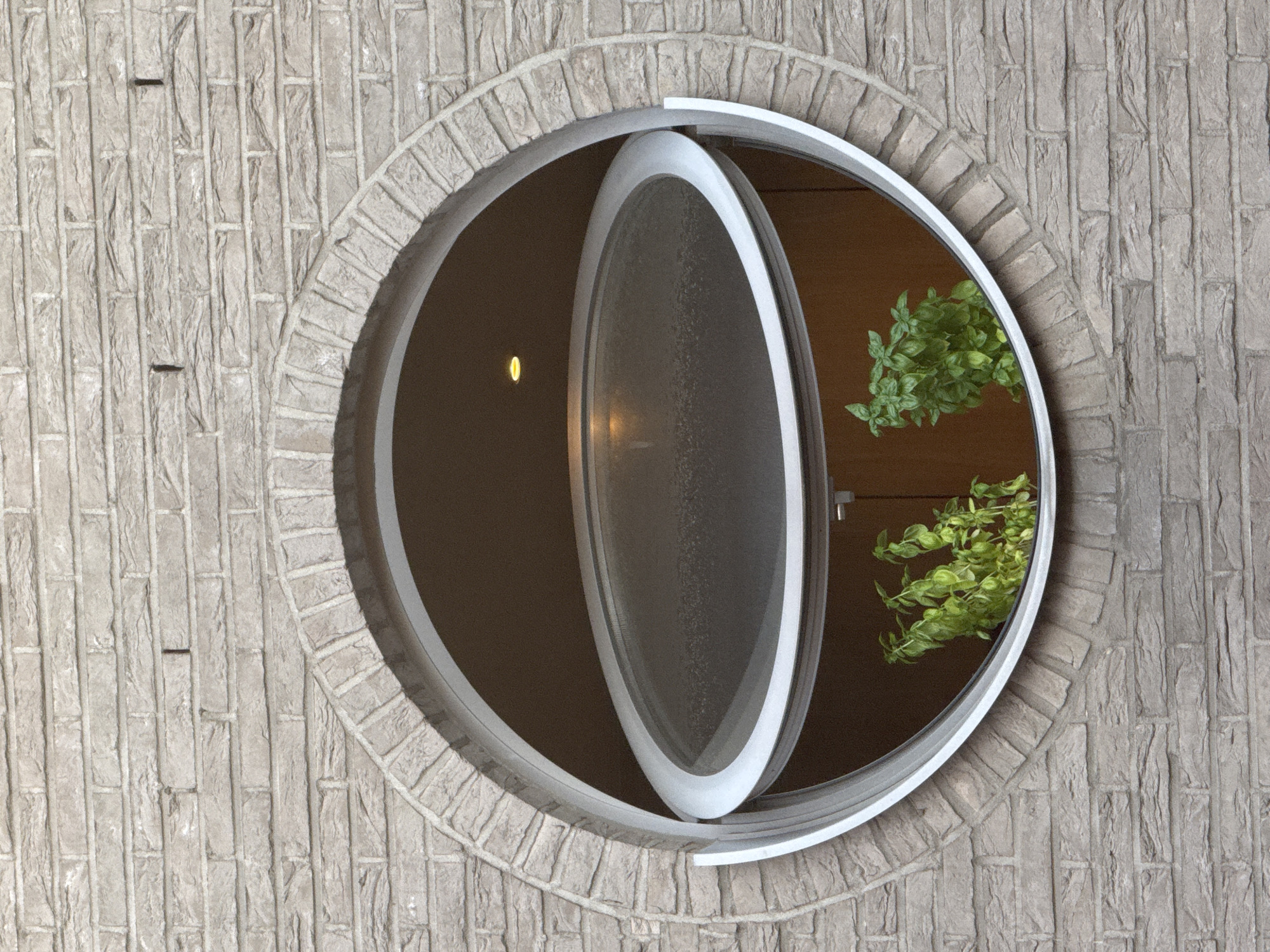 The image shows a close-up of a circular window in a brick building. The window is framed by a light-colored metal trim and has a glass pane that appears to be slightly frosted. The window is partially open, with the lower section tilted outwards. Inside the window, two potted green plants are visible. The brickwork of the building is a light grey color with visible mortar lines. The overall scene suggests an exterior architectural detail. The location is identified as Strombeek-Bever, Belgium.