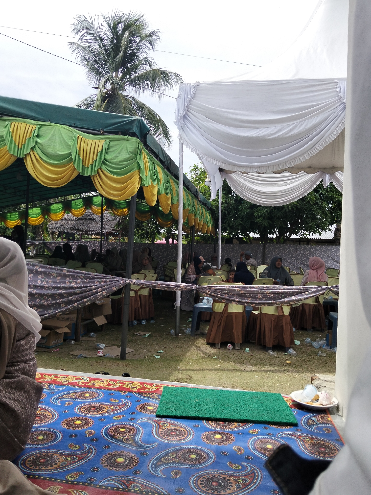 The image depicts an outdoor event, likely a gathering or ceremony, set up under two tents. The tent on the left is green and yellow with decorative draping, while the tent on the right is white with similar draping. Several people, mostly women wearing headscarves, are seated on chairs arranged around tables. The ground is covered with grass and scattered debris, including plastic bottles and cups. In the foreground, a colorful patterned rug is visible, along with a small green mat and a plate with a few items. A palm tree is prominent in the background, and the overall impression is of a casual, community event taking place in a tropical or subtropical environment. The location is identified as Cottrueng, Indonesia.