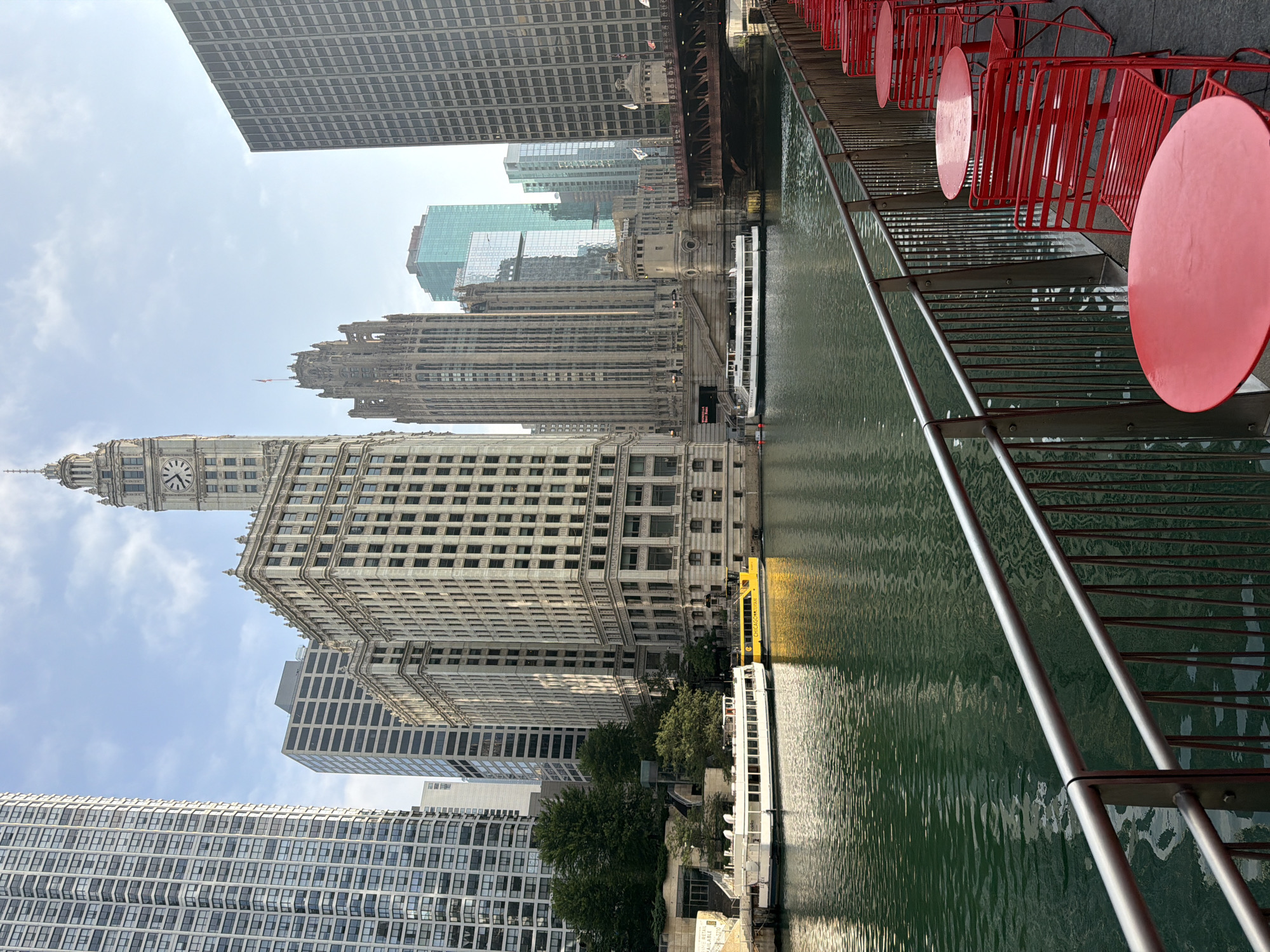The image depicts a view of the Chicago River in Chicago, Illinois, USA. The river occupies the foreground and middle ground, reflecting the surrounding buildings. On the left bank of the river, there are two boats docked: a white tour boat and a yellow water taxi. Behind the boats, a building with the word "CHICAGO" is visible on the yellow taxi. In the background, a series of skyscrapers line the river, with notable structures including a clock tower building, a gothic-style skyscraper, and modern glass buildings. A bridge with red accents spans the river in the mid-ground. In the right foreground, a row of red metal tables and chairs are set up on a paved area next to a metal railing, overlooking the river. The sky is partly cloudy.