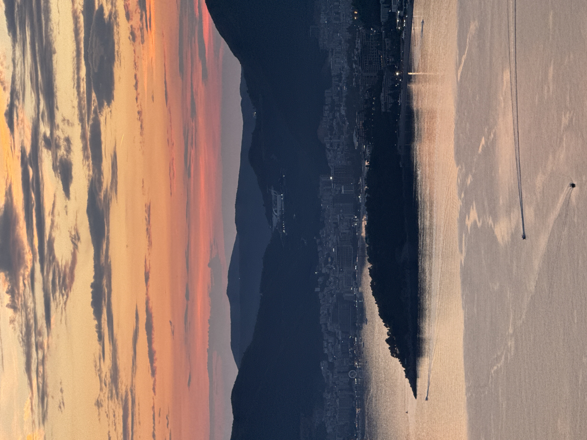 The image captures a panoramic sunset scene overlooking a coastal city. The sky is a gradient of warm oranges and soft pinks, with streaks of dark clouds. Silhouetted against the vibrant sky are mountains that form a dramatic backdrop to a sprawling city. The city lights are beginning to twinkle, indicating dusk. A body of water, likely the sea, reflects the colors of the sunset, creating a shimmering effect. The water appears calm, with a few small boats leaving faint wakes on its surface. The coastline is densely populated with buildings, suggesting a well-developed urban area. The overall mood is serene and picturesque, evoking the beauty of twilight. The location is identified as Čelobrdo, Montenegro, which is known for its stunning coastal views.