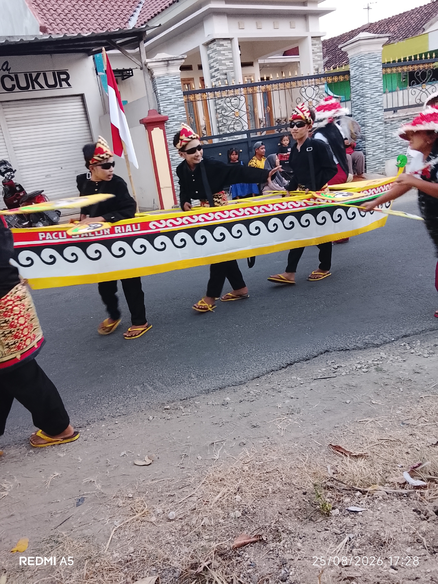 This image captures a procession on a street in Semanding, Indonesia. Three young males, dressed in black shirts, black pants, and traditional headwear adorned with red and white patterns, are carrying a decorative boat-like float. The float is primarily yellow and white, with red trim and a black decorative pattern of swirls. The text "PACU JALUR RIAU" is visible on the side of the float. They are wearing sunglasses and yellow flip-flops. Behind them, other individuals, including women and children, are visible in the background, some observing the procession. A red and white Indonesian flag is displayed prominently on a pole. The street is paved, with a dry, grassy verge on the right. Buildings with tiled roofs are situated behind a metal fence. A motorcycle is parked on the left side of the image. The scene appears to be a cultural event or celebration.