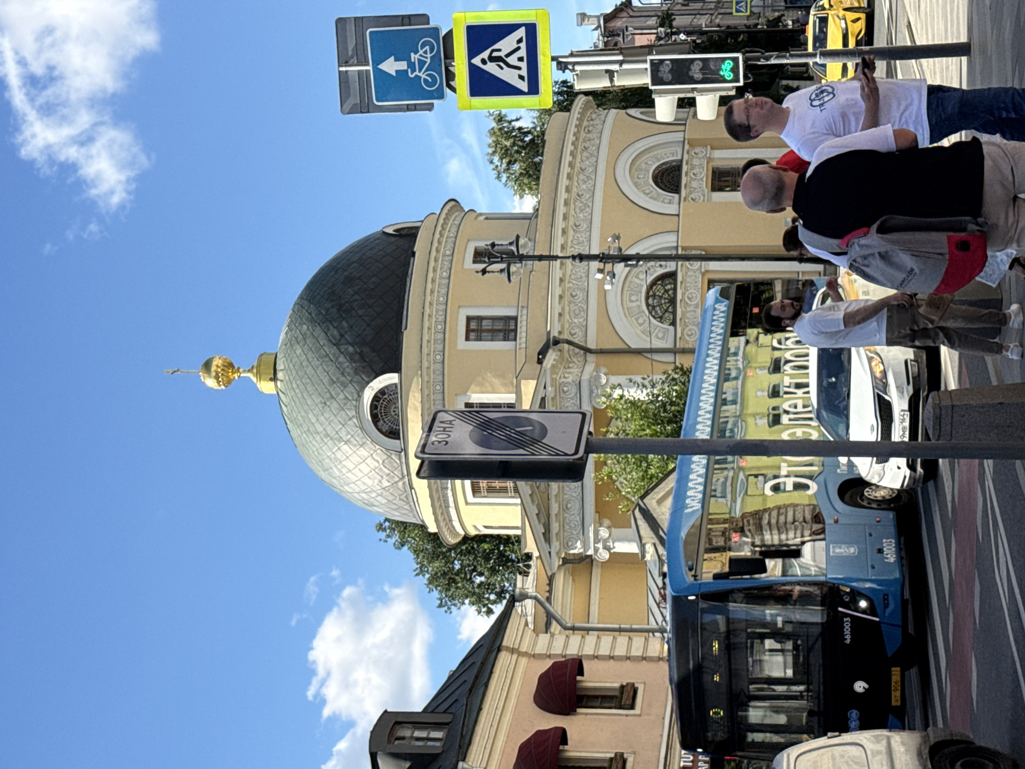 The image captures a sunny day in Moscow, Russia, with a bright blue sky dotted with fluffy white clouds. In the foreground, a blue trolleybus with white decorative trim is stopped at a crosswalk. The trolleybus has the number "461003" displayed on its front and side. To the right of the trolleybus, a white car is also stopped at the crosswalk. Several people are visible. Three men are standing on the sidewalk: one in a white t-shirt and khaki pants, another in a black and white t-shirt with a backpack, and a third in a white t-shirt and jeans. Behind them, a yellow taxi is visible in the distance.
The backdrop of the scene features a light yellow building with architectural details, including columns and arched windows, topped by a dark dome with a golden cross. A street lamp with multiple lights stands in front of the building. Several traffic signs are visible: a blue sign with a bicycle and an upward arrow, a yellow sign with a pedestrian crossing symbol, and a vertical sign with a circle and diagonal lines, indicating a prohibited zone. A traffic light showing a green bicycle symbol is also present. The street appears to be a busy urban intersection with pedestrian and vehicular traffic.