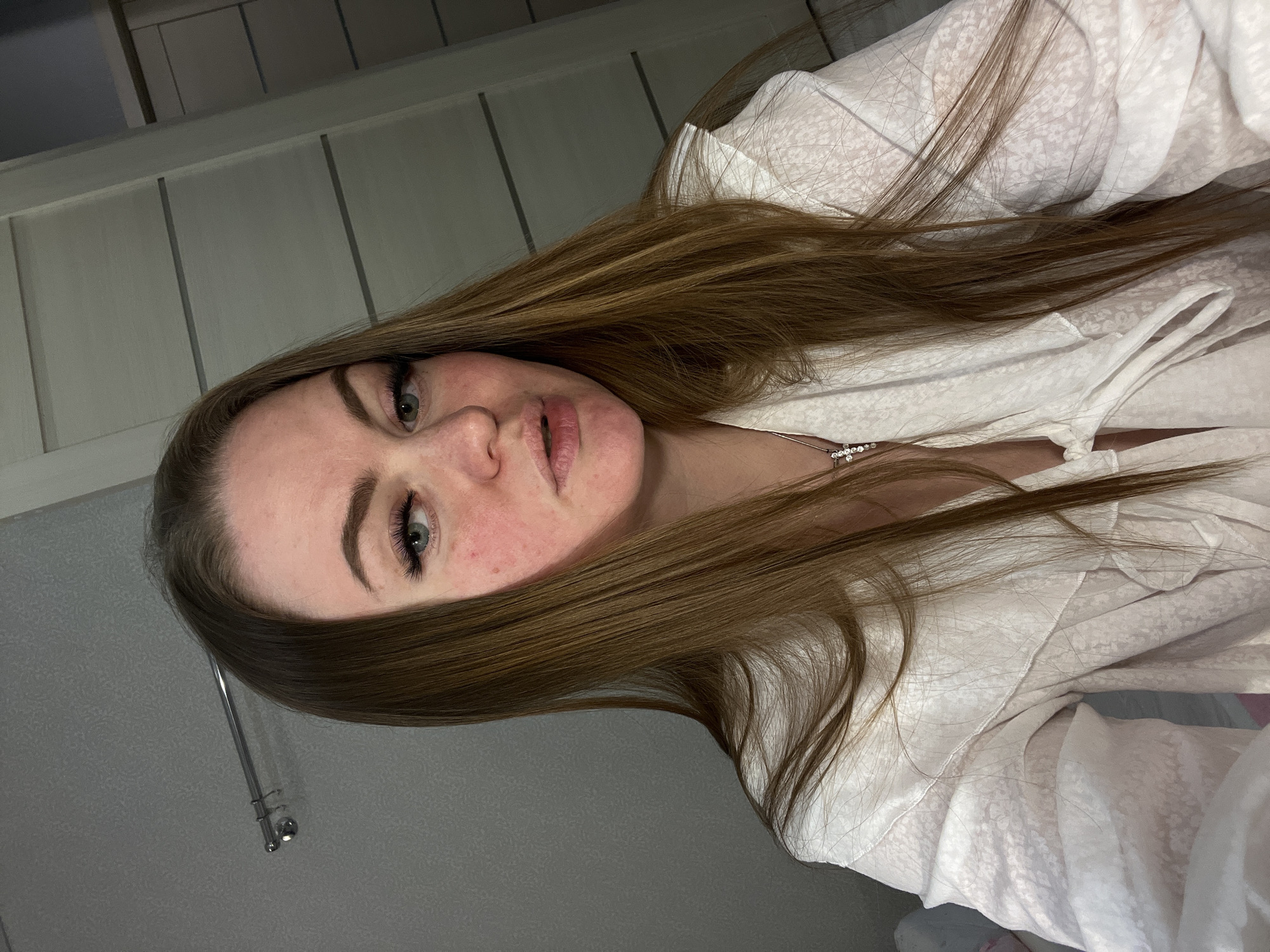 The image is a portrait-style photograph of a young woman taken indoors, appearing to be a selfie due to the upward angle and close proximity. The woman has long, straight, medium brown hair that cascades around her shoulders and chest. She has fair skin with visible freckles and some redness on her cheeks. Her eyes are light (blue or green), and she has dark, prominent eyelashes. Her lips are full and appear naturally colored. She is wearing a light-colored, possibly white or off-white, top with a subtle textured or patterned fabric and a V-neckline. A delicate silver-toned necklace with small, clear gemstones is visible around her neck.
The background shows two distinct wall sections. To her left, there is a light-colored paneled wall, and to her right, a plain grey wall with a metallic, silver-colored rod or fixture attached. The lighting is soft and indirect, suggesting an indoor environment with artificial light, possibly in the evening or a dimly lit room. The woman's expression is neutral and calm, looking directly towards the camera. No text is visible in the image. While the provided location context is Sevastopol, Ukraine, there are no specific visual cues within the image to confirm this geographic setting.