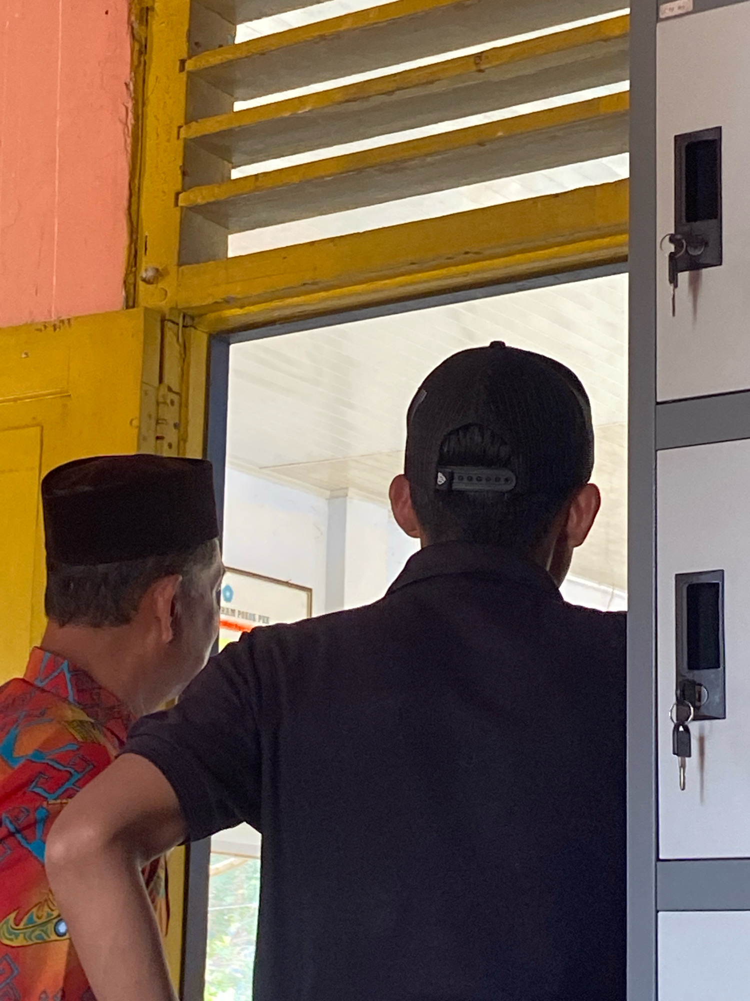 The image depicts two individuals standing in what appears to be a hallway or entryway with lockers to the right and a yellow-painted doorframe with louvers above. The person on the left is wearing a black peci hat and a red shirt with a blue and yellow patterned design. The person on the right is wearing a black t-shirt and a black baseball cap. They are both looking towards the right, with the person on the right facing away from the camera. The lockers are light gray with dark gray keyholes. The environment suggests a public or institutional setting. The image does not provide specific details to confirm the location within Mojo Pahit, Indonesia.